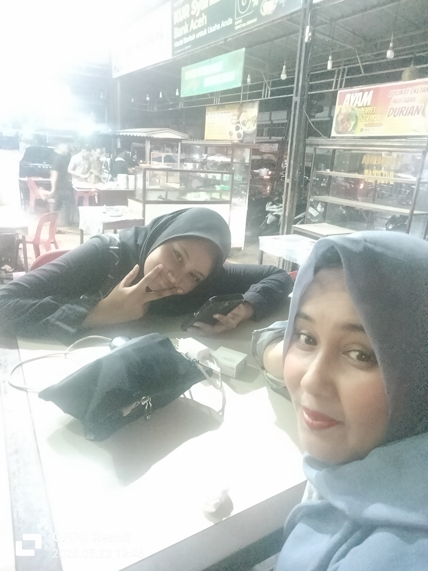 Two women wearing hijabs are seated at a table in what appears to be an outdoor dining area. The woman on the left is wearing a dark hijab and is resting her head on her hand, giving a peace sign gesture. She is holding a smartphone. The woman on the right is wearing a lighter blue hijab and is taking a selfie. A black bag and a white charger are on the table. Behind them are food stalls with signs advertising food such as "Ayam Penyet" (smashed chicken), "Bulukat (Ketan Martabak) Durian," and "Nasi Soto" (soto rice). A sign also shows "KUR Syariah Bank Aceh" as the area is located in Meukokuthang, Indonesia. The background shows other diners and food preparation areas. The timestamp "2026.05.23 19:46" indicates the image capture date.