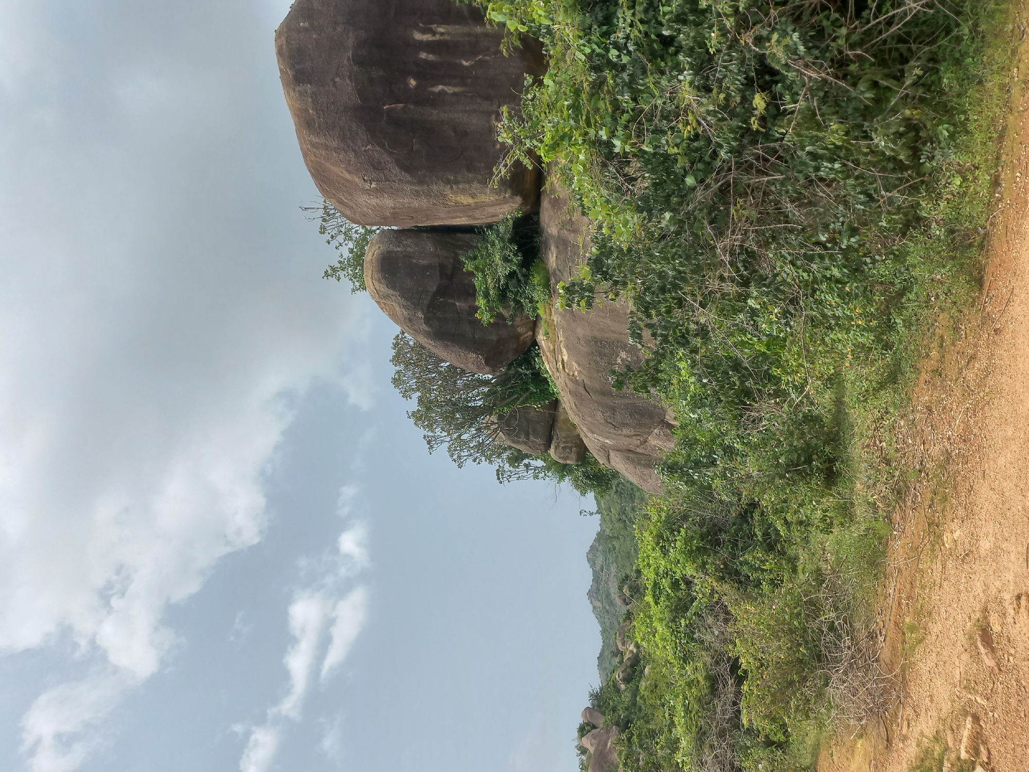 The image shows a natural landscape dominated by large boulders and lush green vegetation under a cloudy sky. The foreground features a dirt path with some scattered dry branches. In the mid-ground, a cluster of massive, rounded, brownish boulders are prominent, partially covered by green shrubs and trees. The background consists of rolling hills covered in dense vegetation, characteristic of the outskirts of Jos, Nigeria. The overall scene depicts a rugged, undeveloped natural area.