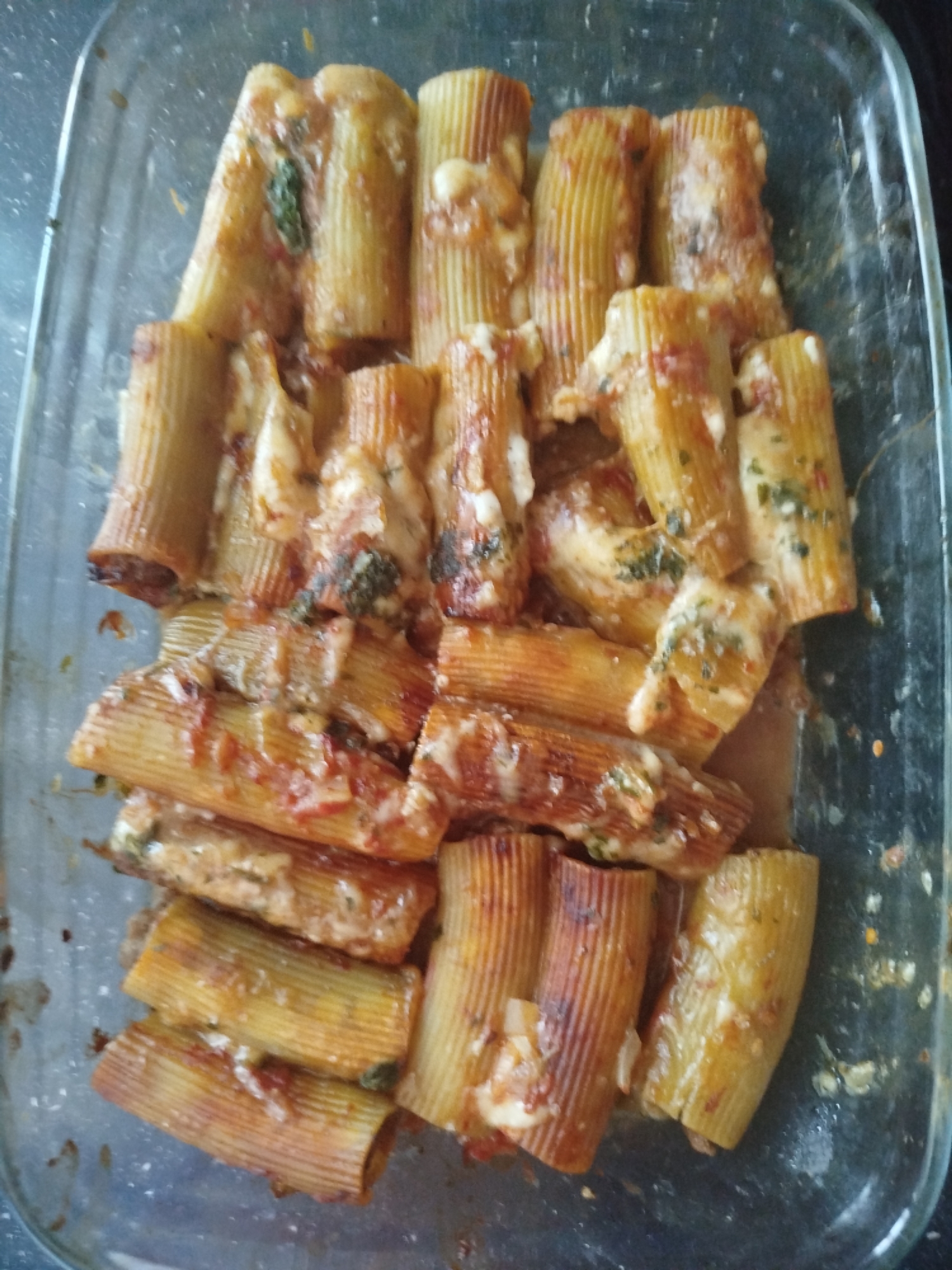 A close-up, top-down shot shows a glass baking dish filled with rigatoni pasta tubes. The pasta is covered in a rich red sauce, melted cheese, and what appears to be herbs, possibly basil or parsley. The rigatoni is stuffed and baked, with some tubes showing a browned, slightly caramelized exterior. The dish is filled to the brim, with the pasta overlapping and forming layers. The glass dish has a slight sheen and some residue is visible on its sides. The lighting is somewhat dim, creating shadows that highlight the textures of the pasta and sauce. There are no people or external environmental elements visible. The scene is limited to the food within the baking dish.