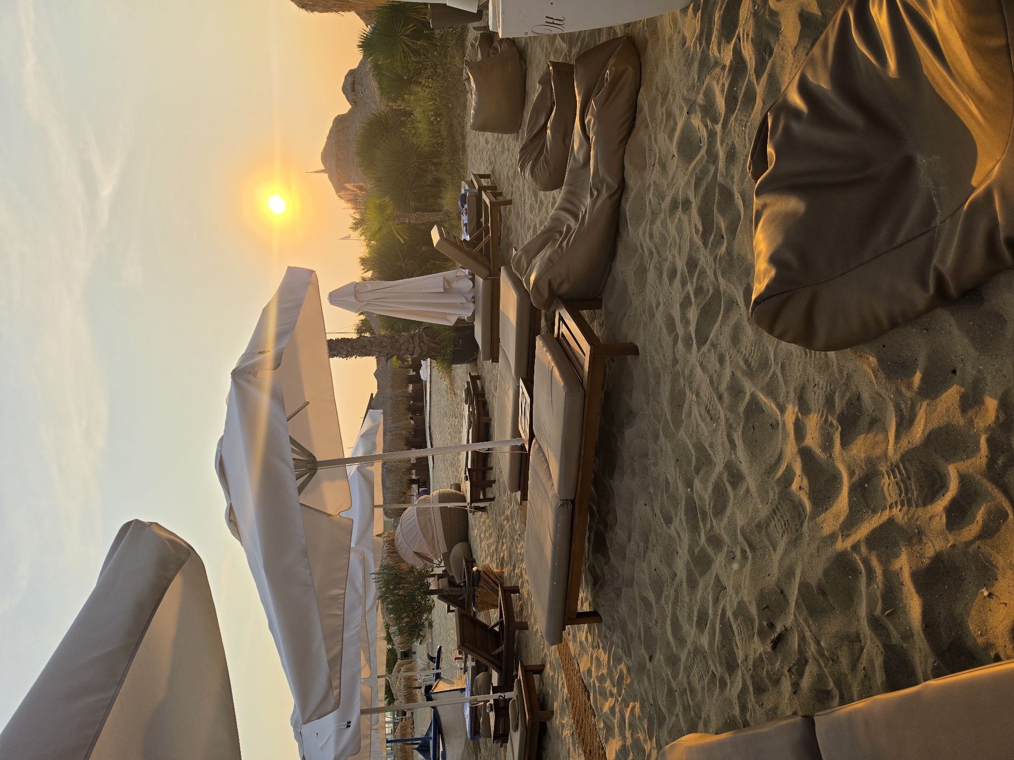 The image depicts a serene beach scene at sunset in Sveti Vlas, Bulgaria. The sandy beach is furnished with numerous lounge chairs and large, beanbag-style seating, all arranged under white parasols that offer shade. The setting sun casts a warm, golden glow across the sand and illuminates the sky with soft orange and yellow hues. In the background, palm trees and thatched-roof structures add to the tropical ambiance. The scene is devoid of people, suggesting a tranquil, late-day atmosphere. The overall impression is one of relaxation and peaceful enjoyment of the coastal environment.