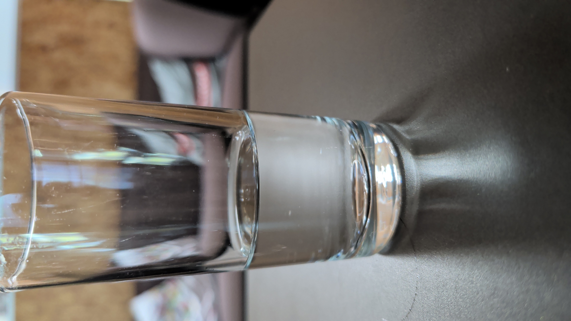 The media file is a photograph of a clear glass tumbler filled with a clear liquid, likely water. The glass is positioned horizontally and rests on a dark, textured surface, possibly a table. The tumbler is cylindrical with a thick base and a rim that appears slightly thicker than the body of the glass. The liquid inside fills approximately half of the glass, and the surface of the liquid is smooth.
To the left of the glass, a blurred background shows what appears to be furniture, with wooden tones and hints of fabric in muted colors. The lighting in the image creates a shadow of the glass on the dark surface to its right, indicating a light source from the left or above. The scene is likely indoors, given the controlled lighting and background elements. There is no visible text in the image. The overall impression is one of simplicity and focus on the object itself, with no discernible activity or specific event taking place. The image has a slightly grainy texture, suggesting it was taken with a digital camera or smartphone. The orientation of the photograph is unusual, with the glass laid on its side.