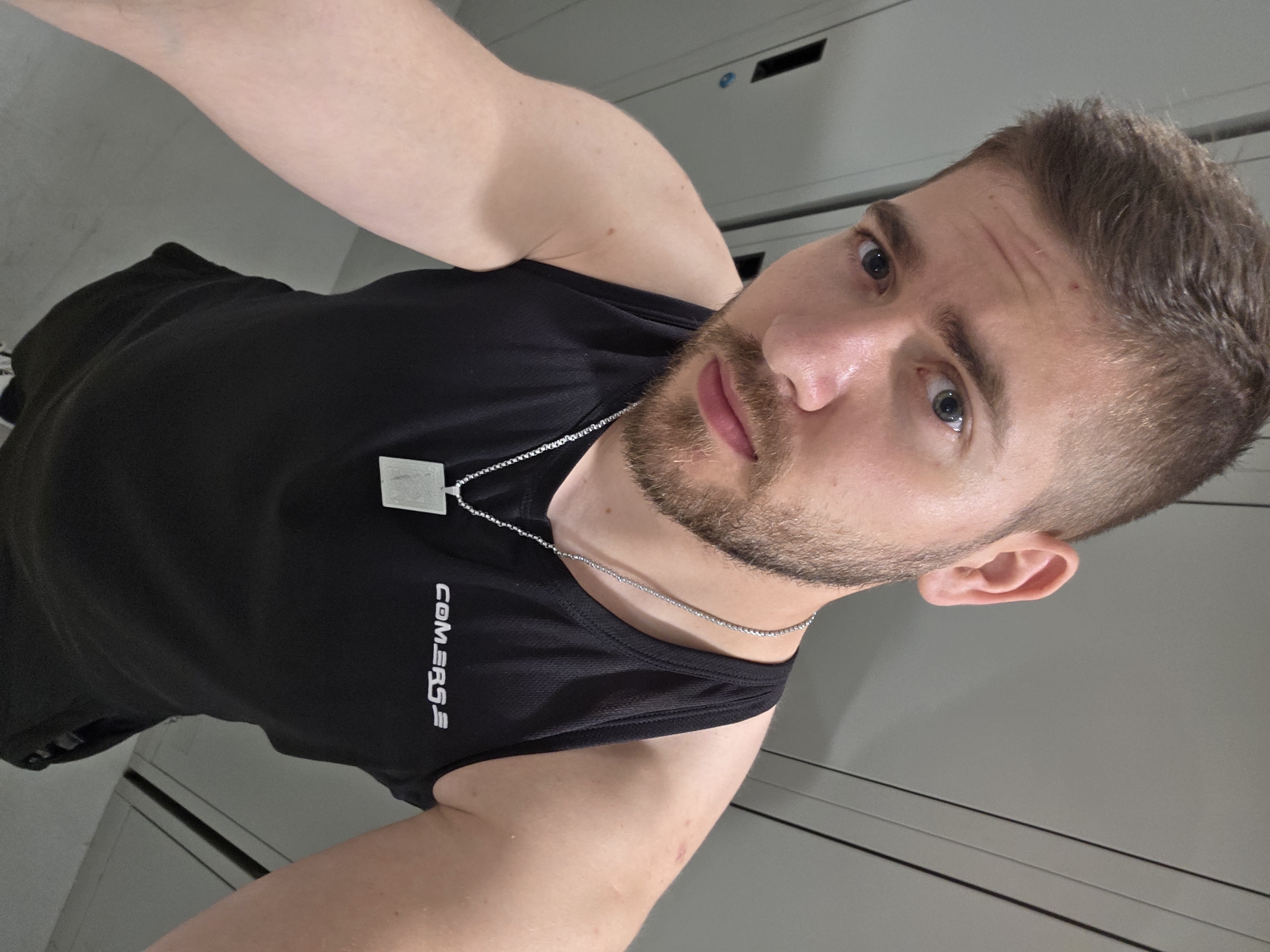 A young man with light skin, short brown hair, and a trimmed beard or stubble is the central subject of this close-up selfie. He has blue eyes and a neutral expression, looking directly at the camera. He is wearing a black sleeveless athletic top, upon which the brand "COMERSG" is printed vertically in white text on the left side of his chest. A thin silver chain with a square pendant hangs around his neck. The image is taken from a slightly tilted and upward angle, showcasing his upper body. The background consists of plain, light gray or white surfaces with subtle horizontal lines, strongly suggesting an indoor environment such as a locker room or changing area. The lighting is even, typical of indoor settings. No explicit visual details within the image indicate the specified location context of Plovdiv, Bulgaria.