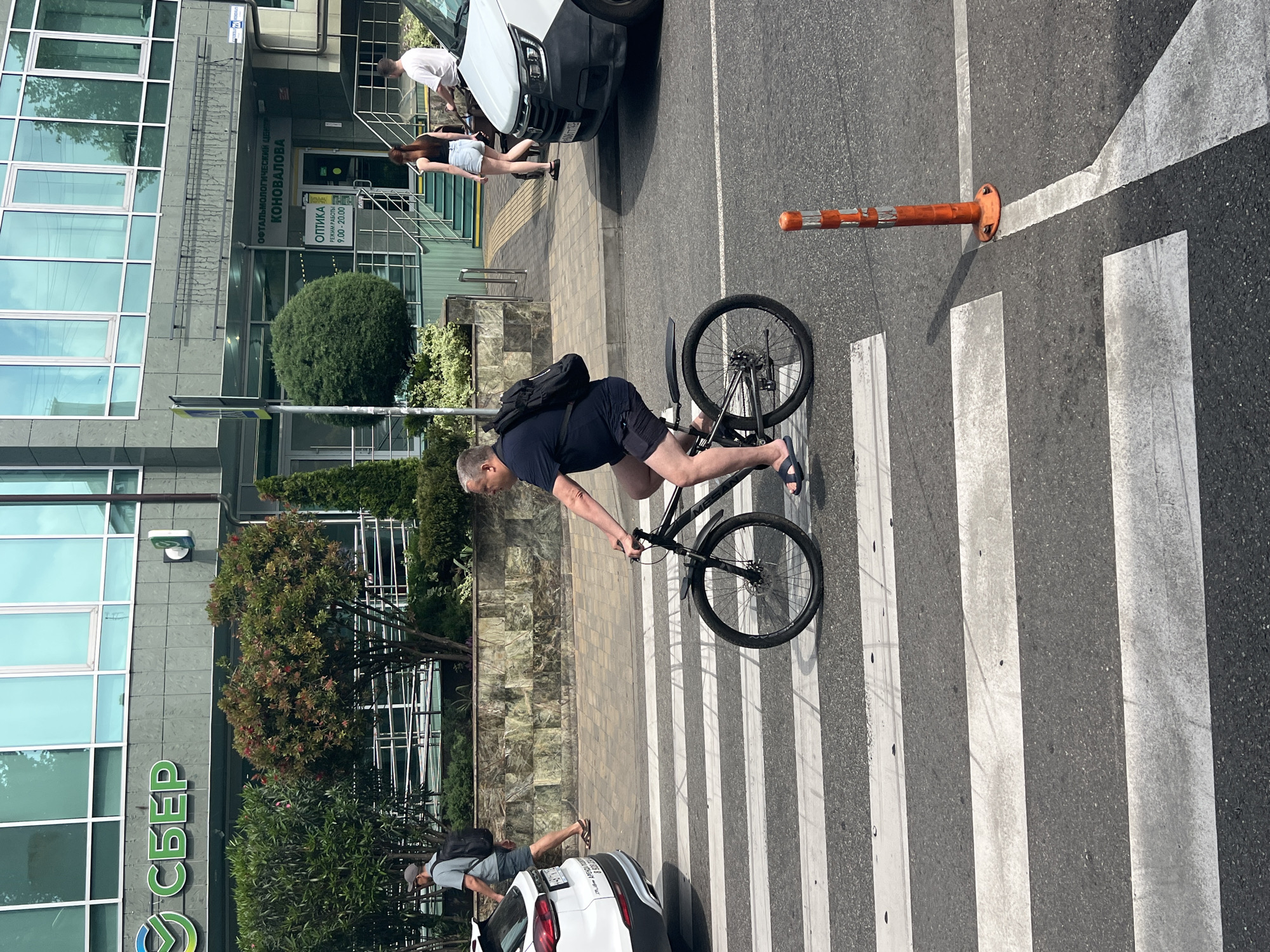 In Sochi, Russia, on a sunny day, a man is cycling across a pedestrian crossing. He is wearing a dark blue t-shirt, dark shorts, and blue sandals, with a black backpack slung over his shoulders. The bicycle he is riding is dark in color. The pedestrian crossing has white stripes on a dark asphalt road.
Behind the cyclist, to the left, a white car is parked, and a man in a grey t-shirt and shorts, wearing a cap and carrying a backpack, is walking on the sidewalk. To the right of the pedestrian crossing, a white van is visible.
The background features a modern building with glass windows. A prominent sign with green letters reads "СБЕР" (Sberbank), indicating the presence of a financial institution. Another sign above an entrance reads "ОФТАЛЬМОЛОГИЧЕСКИЙ ЦЕНТР КОНОВАЛОВА" (Ophthalmological Center Konovalova) and "ОПТИКА" (Optics) with operating hours. Green bushes and trees are planted in front of the building, adding to the urban landscape. A bright orange traffic cone with white reflective bands stands near the pedestrian crossing.