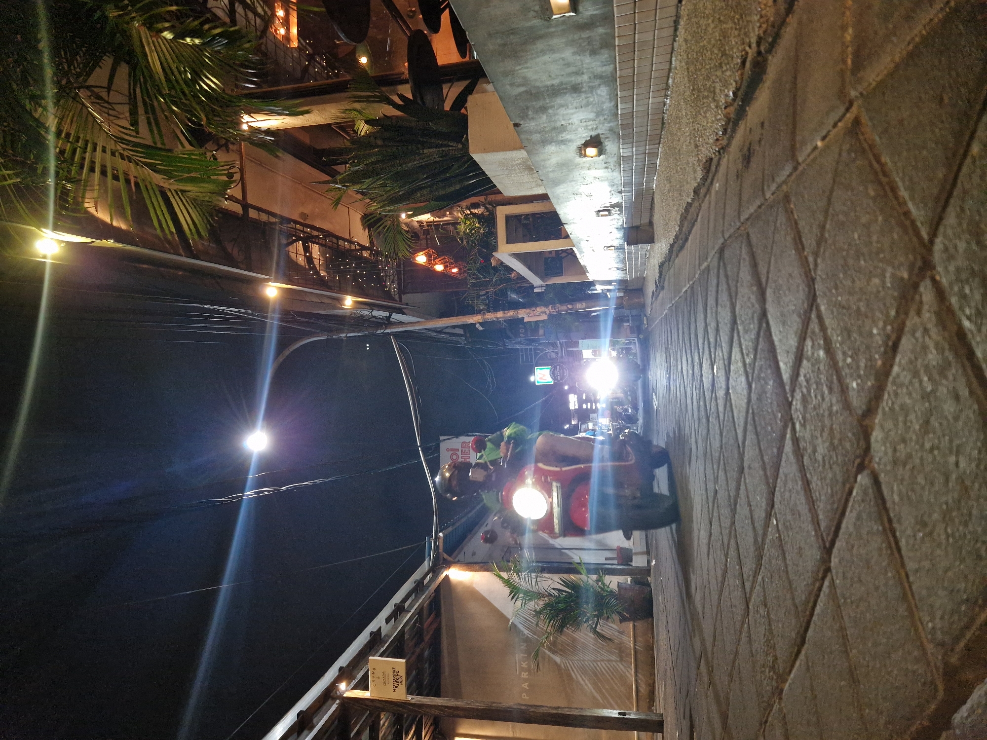 A slightly rotated shot of a Kuta, Indonesia street at night shows a red scooter, with the rider and passenger wearing helmets, driving up the right side of the road, close to the sidewalk. The sidewalk is paved in a dark gray patterned tile. Palm trees are prominent on the left, and a building with multiple levels and lights is visible. The street is lit by streetlights. A scooter in the background has very bright headlights. The scooter's headlights and taillights are illuminated. A sign is visible on the left side of the road.