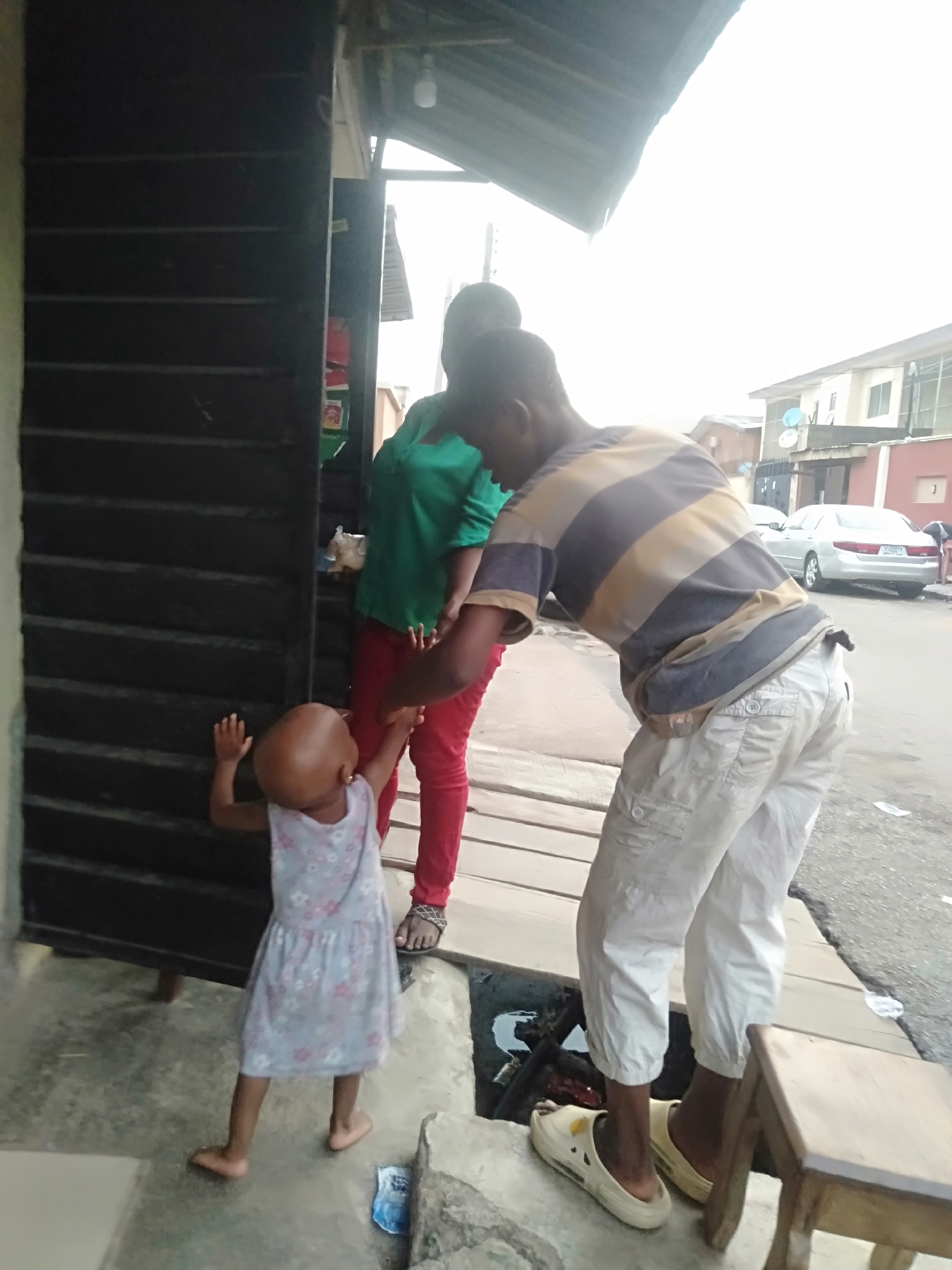 The image depicts a street scene in Shangisha, Nigeria. A small child in a floral dress stands barefoot near the entrance of a dark-colored building with louvered doors. A woman in a green top and red pants stands near the child. A man in a striped shirt and white pants is bent over, his arm extended towards the child. The man is wearing white shoes and standing on a concrete ledge near a drainage ditch that is partially covered with wooden planks. A small wooden stool is beside him. In the background, a silver car is parked on the street. A multi-story building is visible across the street, with satellite dishes mounted on its roof. Debris is scattered on the street. The lighting suggests an overcast day.
