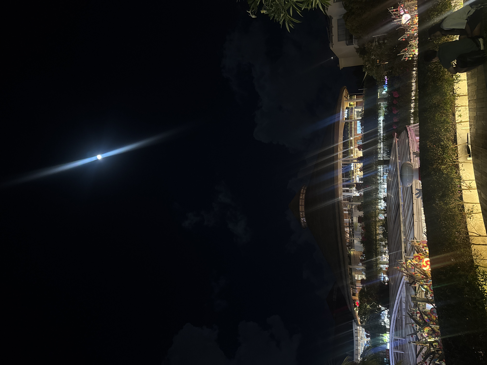 This image captures a night scene in Kuta, Indonesia. The dark sky is illuminated by a bright full moon, casting a luminous beam. The foreground features a lush green hedge, with a stone wall visible to the right. Beyond the hedge, a row of buildings with lit windows suggests a commercial or residential area. In the mid-ground, a decorative display of colorful lollipops on sticks adds a festive touch. The scene appears to be an outdoor area, possibly a promenade or a walkway lined with greenery and illuminated by artificial lights. The presence of the moon indicates it is nighttime. There is no discernible activity taking place, and no clear emotional tone is conveyed. The word "PLAYHOUSE" is visible in neon lights above the lollipop display.