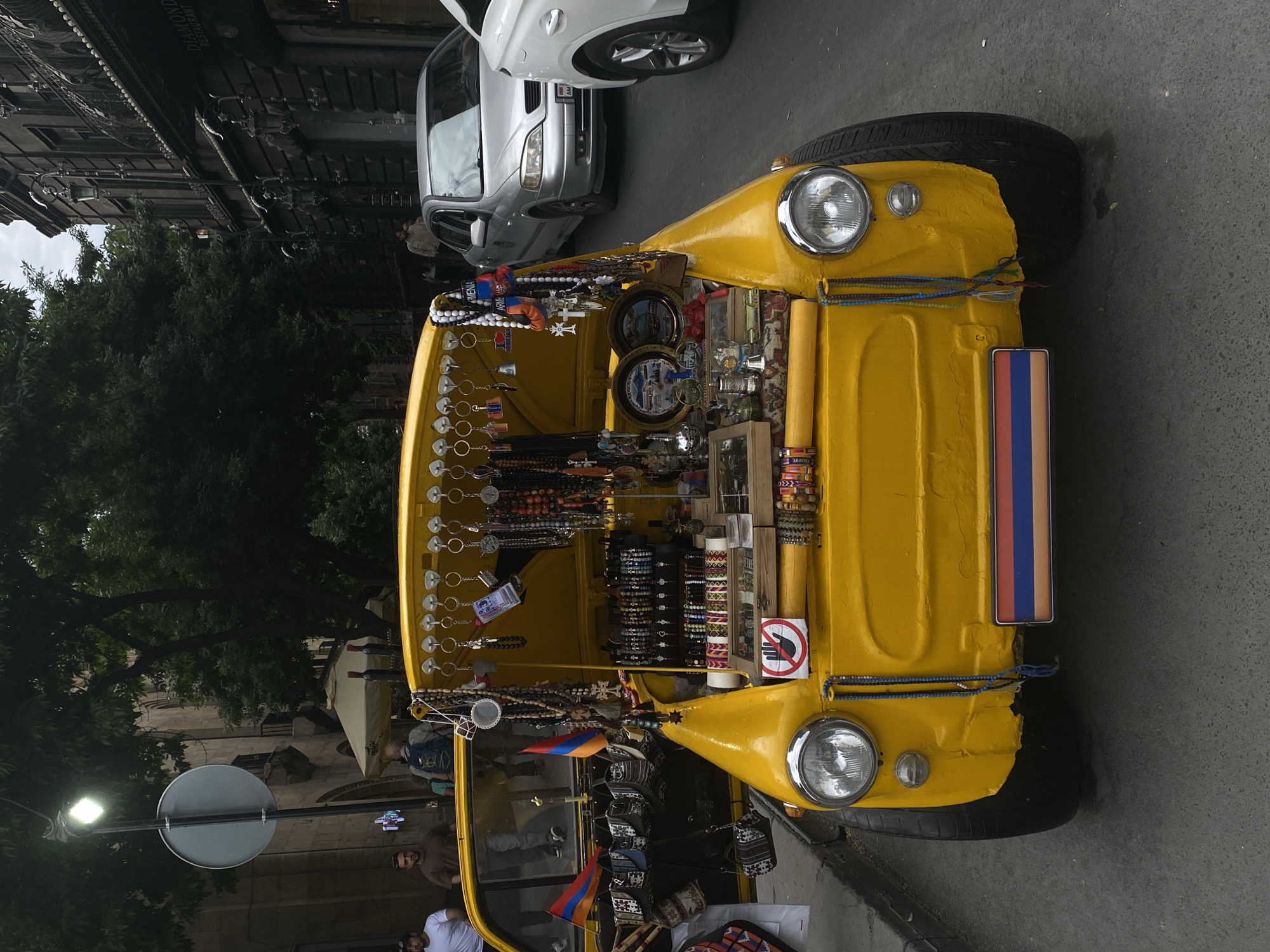 This image captures a vibrant yellow three-wheeled vehicle, likely a modified auto-rickshaw, transformed into a mobile souvenir shop, parked on a paved street in Yerevan, Armenia. The vehicle is brightly painted and extensively decorated with various merchandise.
The main subject is the yellow vehicle, which displays a wide array of souvenirs including beaded necklaces and bracelets, keychains, and small decorative plates or trays featuring imagery. Many items clearly incorporate the colors of the Armenian flag or have "Armenia" inscribed on them. Small rectangular Armenian flags (red, blue, orange) are prominently displayed both as a sign on the front of the vehicle and as miniature flags within the display area. A red circular sign with a hand symbol and a diagonal line, indicating "Do Not Touch," is visible on the side of the vehicle's display.
In the background, a silver sedan is parked, its license plate showing "AM," which signifies Armenia. Further back, old-style buildings with intricate facades line the street, with one partially visible sign reading "DIAMOND RESTAURANT." Lush green trees provide shade and visual depth. Several people are scattered in the background, appearing to be pedestrians or onlookers.
The scene takes place during the daytime, under what appears to be an overcast sky, contributing to soft, even lighting. A street light and the reverse side of a round traffic sign are visible on the left. The overall impression is a lively, informal street vending operation typical of a city setting.