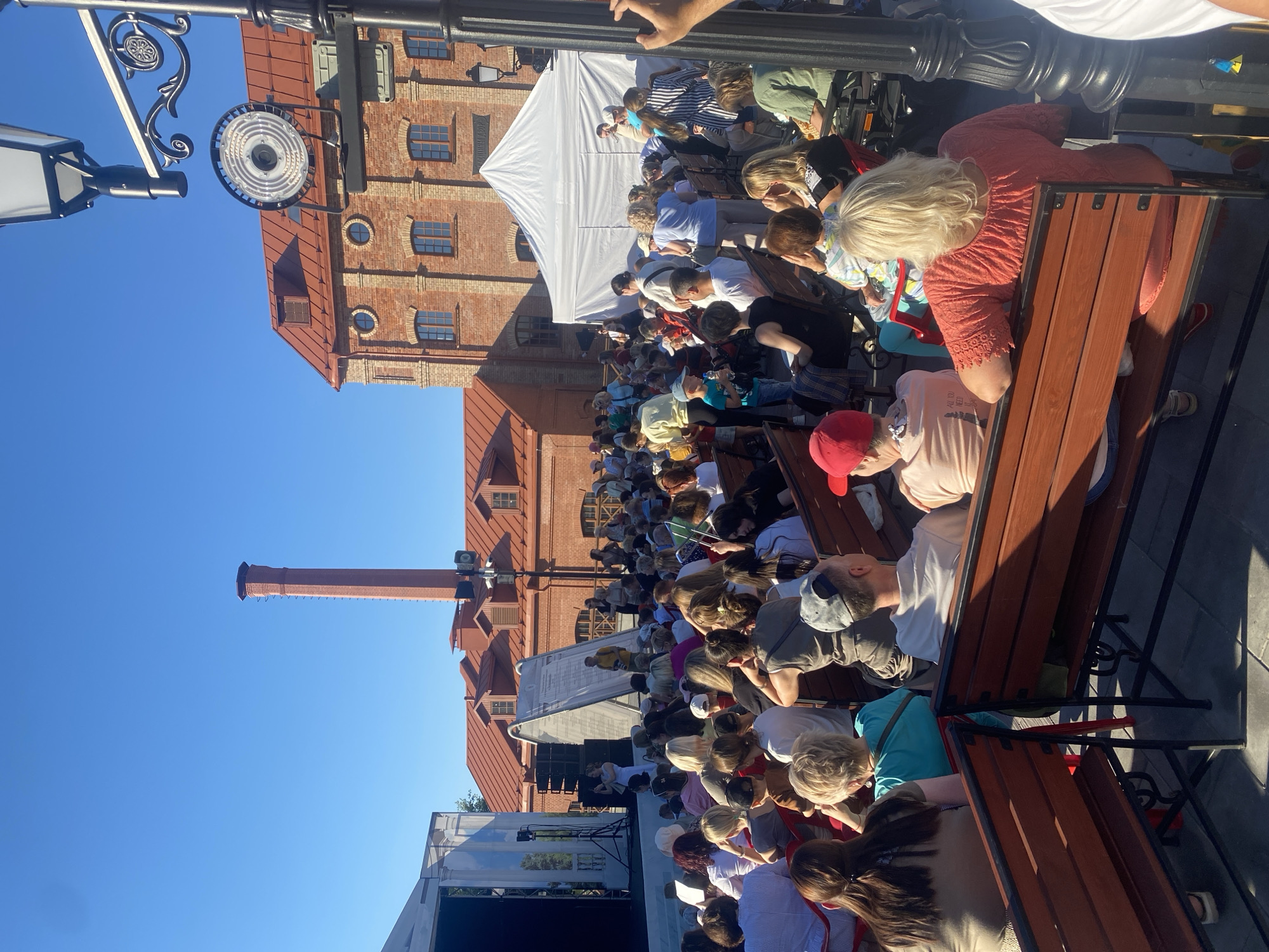 The image depicts an outdoor gathering of people seated on benches, facing a stage area with a white tent and a brick building in the background. The building features a red tiled roof, arched windows, and a tall brick chimney. A street lamp with ornate details is visible in the foreground. The sky is clear and blue. The event appears to be an outdoor concert or performance, possibly part of a festival, taking place in the Upper Town district of Minsk, Belarus.