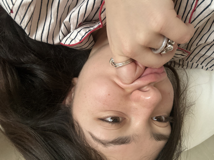 This image depicts a close-up of a young adult female, positioned as if reclining indoors in the city of Porto, Portugal. She has dark, wavy hair, brown eyes, and fair skin with visible freckles. Her left hand is raised to her face, with the index and middle fingers pressing into the right corner of her mouth, causing her lips to pucker and her right cheek to indent. Her gaze is directed slightly upwards and to the viewer's right, conveying a playful expression. She is wearing a light-colored, vertically striped pajama top with dark stripes and red piping. Two silver-toned rings are present on her left hand: a prominent textured ring on her index finger and a smoother ring on her middle finger. The background is an out-of-focus light-colored surface, consistent with a pillow or bedding.