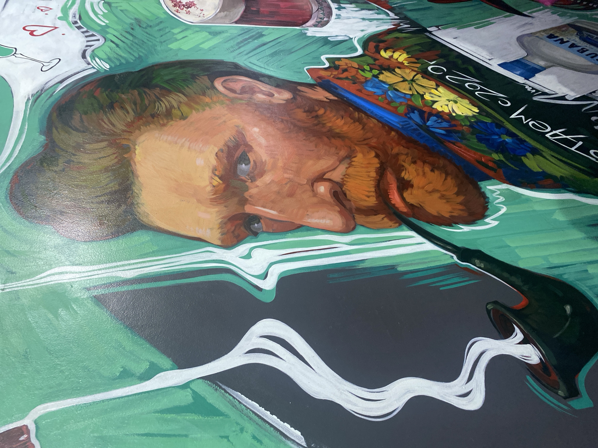 The image depicts a painted mural. A man's head is prominently featured on the left side of the frame, oriented sideways. The head is painted in tones of brown, orange, and green. A bottle is depicted on the lower right with white lines emerging from it. Above the man's head are painted images of a heart, a glass, and food in containers. Floral designs and text appear above the man's head. The dominant colors in the rest of the mural are shades of green, gray, and white. The mural is located in Krasnoyarsk, Russia.