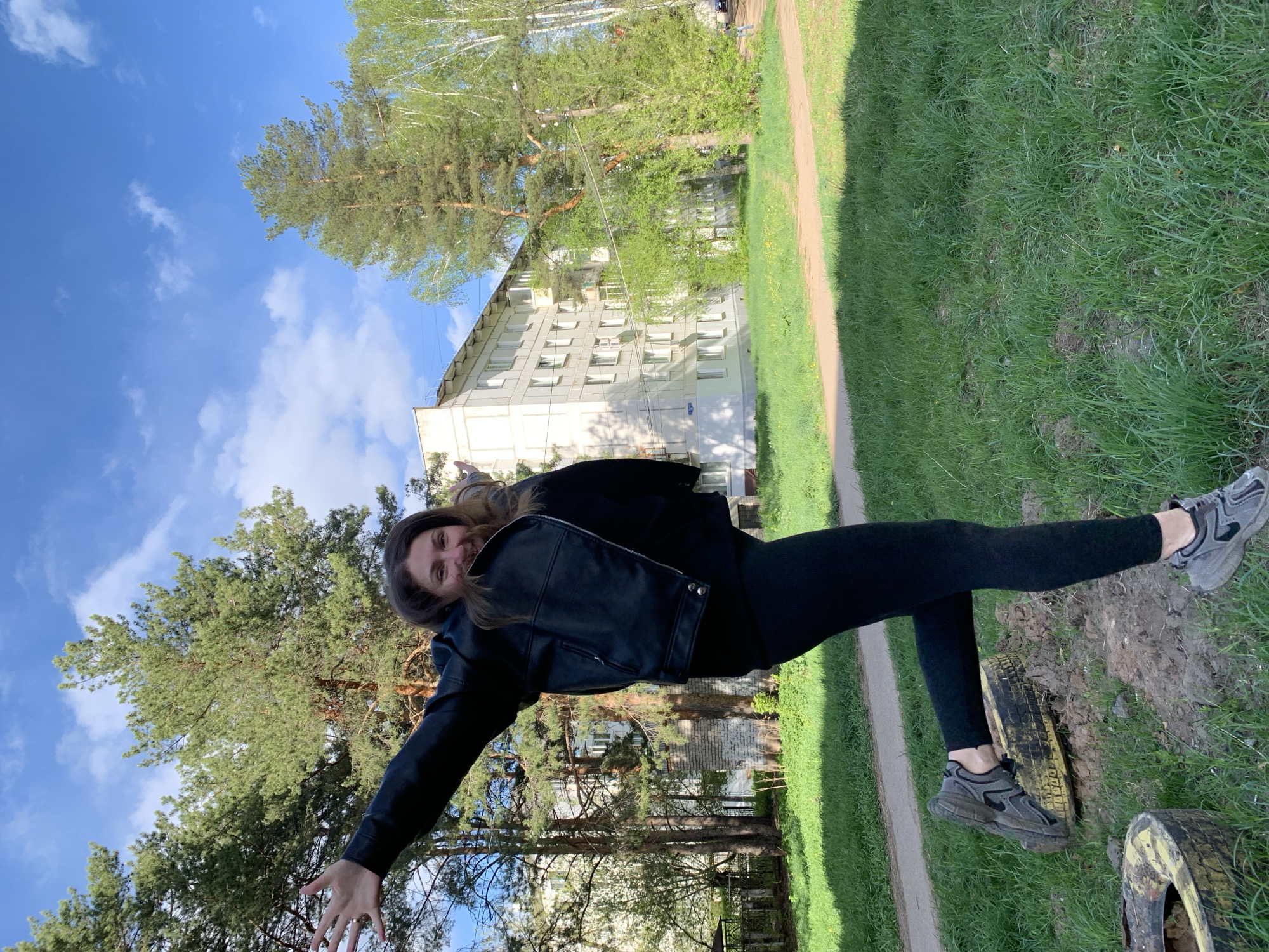 A medium shot photograph taken in the city of Chaikovsky, Russia, captures a woman standing with her legs resting on two tires sunken in the ground, located along a grassy field adjacent to a sandy path. The woman is dressed in black leggings, a black jacket, and gray athletic shoes. She extends one arm towards the camera and smiles, with a building and trees visible in the background beneath a partly cloudy blue sky.