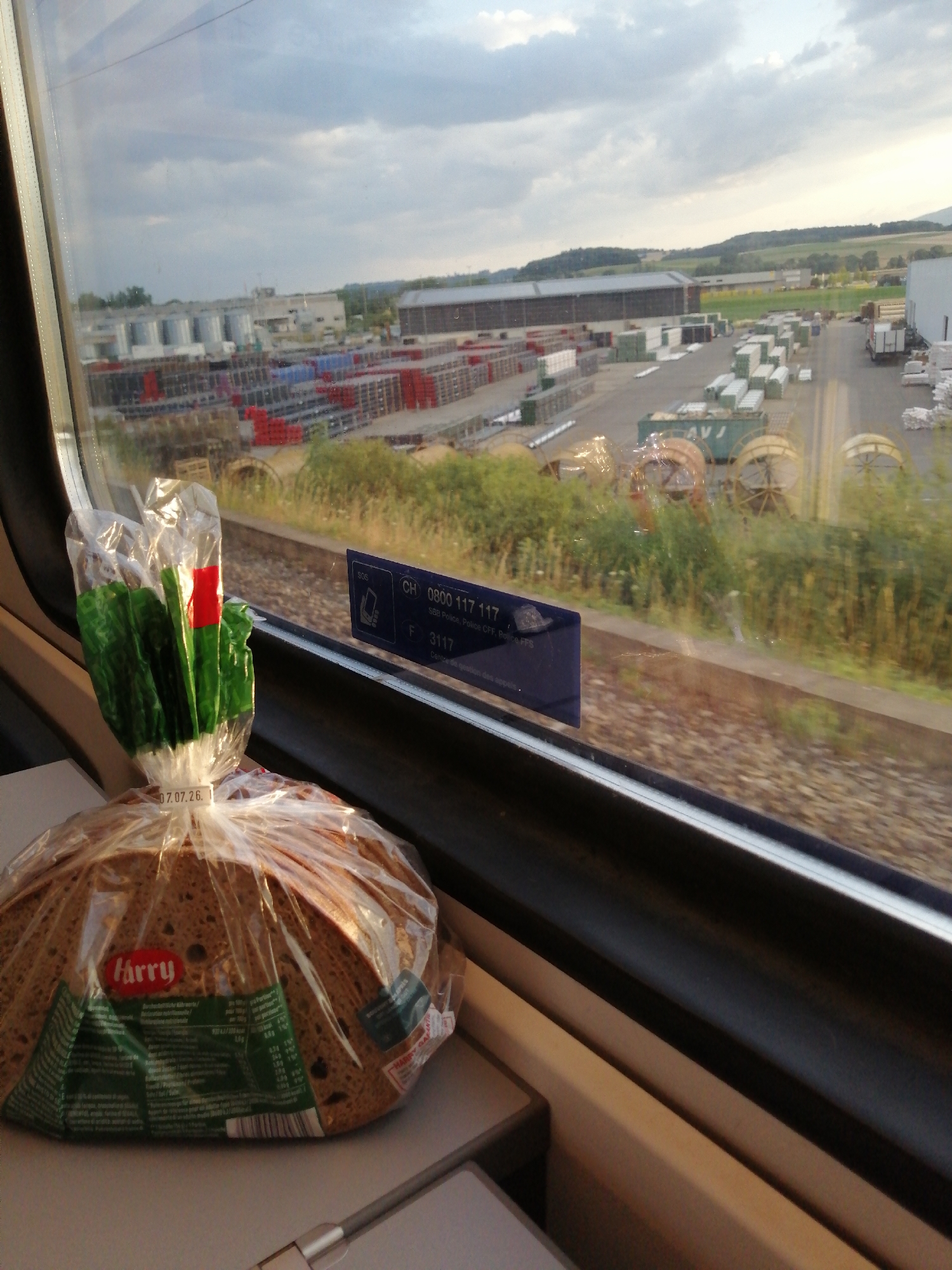 The image is a close-up shot from inside a train, looking out the window. In the foreground, a loaf of bread wrapped in clear plastic is on a tray table. The bread has a label with the brand "harry" and nutritional information. A plastic bag with green leafy vegetables and a red tag is also visible, attached to the bread packaging.
Outside the window, a train track runs parallel to the scene. Beyond the tracks, there is a large industrial area with multiple buildings, stacked pallets of goods, and what appear to be large spools or reels. The landscape in the background shows rolling hills and fields, suggesting a semi-rural or suburban setting. The sky is partly cloudy. A blue sticker with "SBB Police, Police CFF, Police FPS" and a phone number is affixed to the window. The date "07.07.26." is visible on the bread packaging. The location is identified as Eclépens, Switzerland.