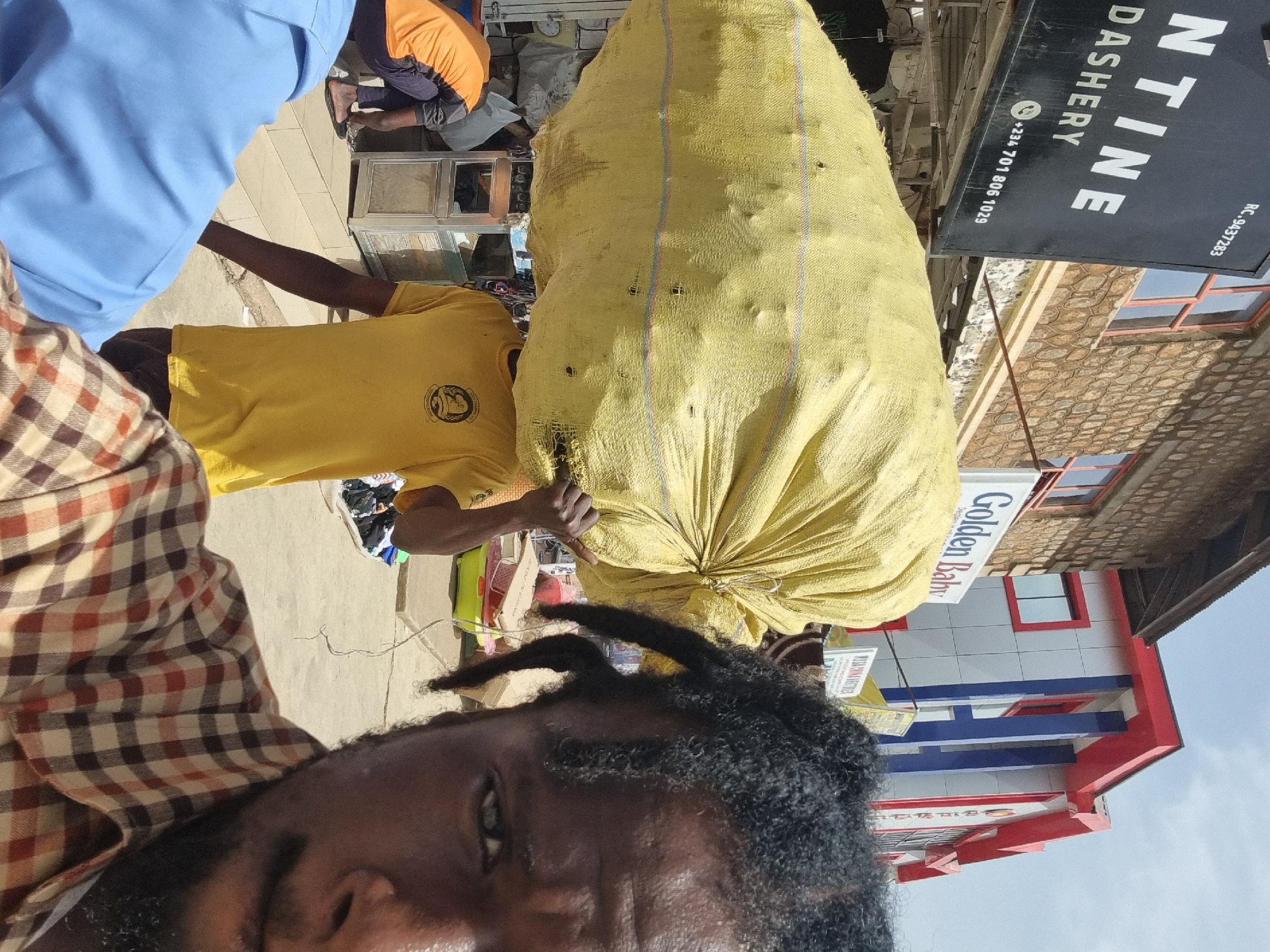 The image, taken in Jos, Nigeria, shows a man carrying a large yellow sack on his shoulder. The man is wearing a yellow t-shirt. A second man, wearing a plaid shirt, is in the foreground and appears to be taking the picture. The background includes storefronts and other people. Storefronts visible include one for "NTINE DASHERY" with the phone number +234 701 806 1029 and registration number RC-9437283. Another business is visible with the name "Golden Baby." The sky is visible in the background.