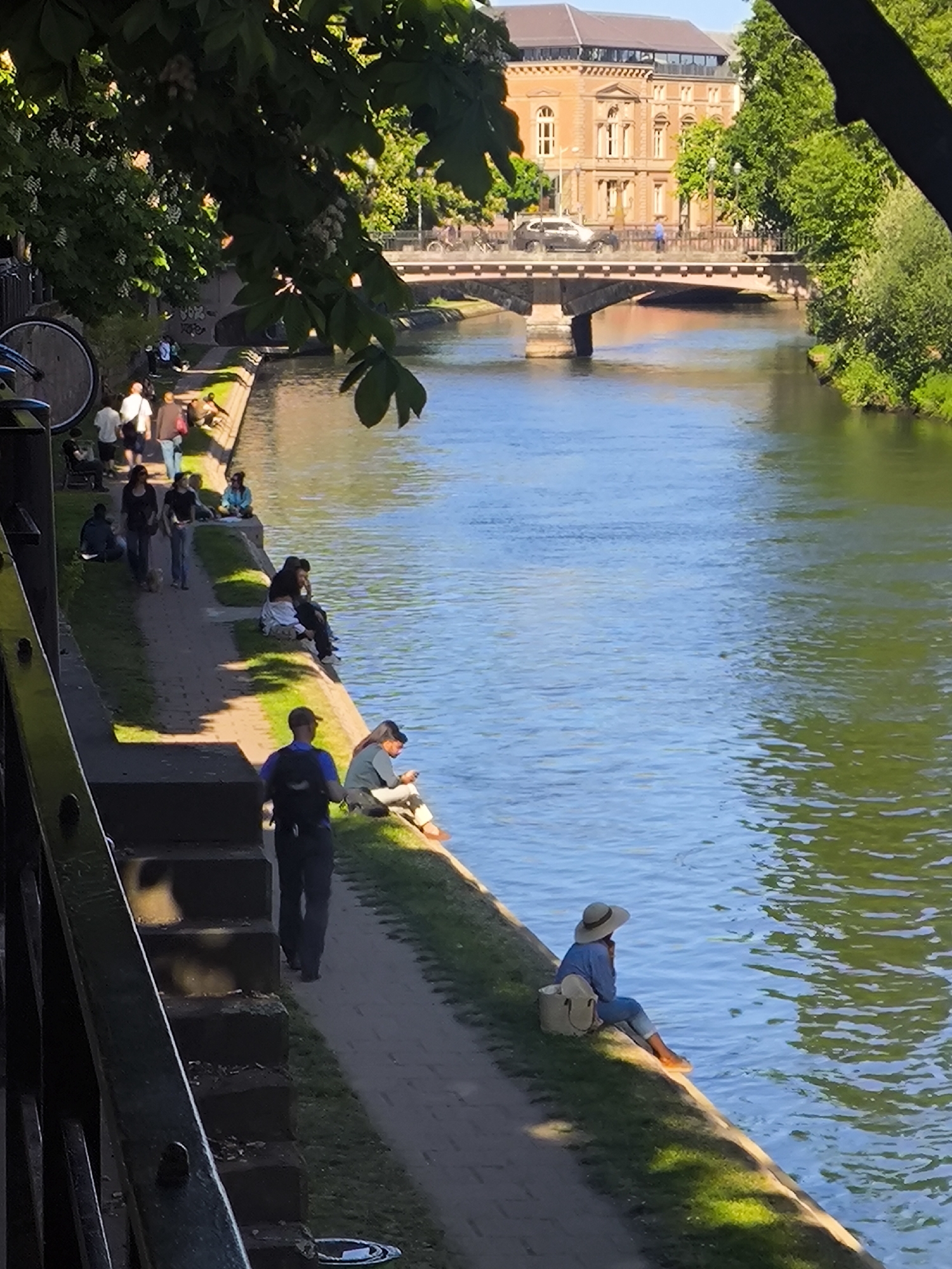 The photograph captures a vibrant scene along the Ill River in Strasbourg, France. The sunlit river is the central focus, reflecting the clear sky. Along the riverbank, a paved pathway is populated with people engaged in various activities. Some are strolling, while others are seated near the river's edge, possibly enjoying the scenery or engaging in personal activities such as reading. In the foreground, a man in a uniform walks along the path. Across the river, a bridge is visible, with a light-colored car and pedestrians crossing. In the background, there's a grand, classical-style building with red brick and architectural details. The scene is framed by greenery, including trees and foliage, creating a natural and serene setting within the urban landscape. The composition suggests a mix of leisure, recreation, and daily life taking place in a picturesque environment.
