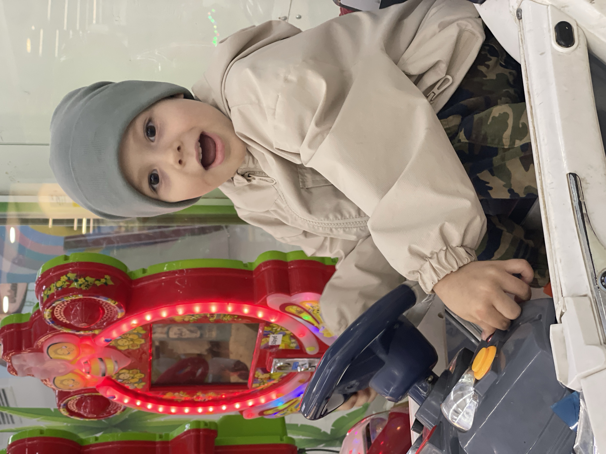 An excited young child is captured playing an arcade driving game in an indoor entertainment setting, likely an arcade or play area. The child, with light skin and dark eyes, sports a wide-open mouth that suggests surprise or delight. They are wearing a grey beanie and a light beige or tan hooded jacket over camouflage-patterned pants.
The child's hands are actively engaged with the game, gripping a blue steering wheel, and their right hand is positioned near an orange button on the game console. The immediate foreground shows parts of the white game machine, emphasizing the immersive experience. In the background, a brightly lit red and green arcade game machine, adorned with glowing red lights and yellow character designs resembling bees, adds to the vibrant atmosphere of the scene. A small, yellow sticker on this background game appears to have indistinct text, possibly "9/096". The setting is characterized by artificial lighting, reflecting off glass surfaces, typical of an indoor amusement venue. The overall mood is one of playful energy and childhood wonder.