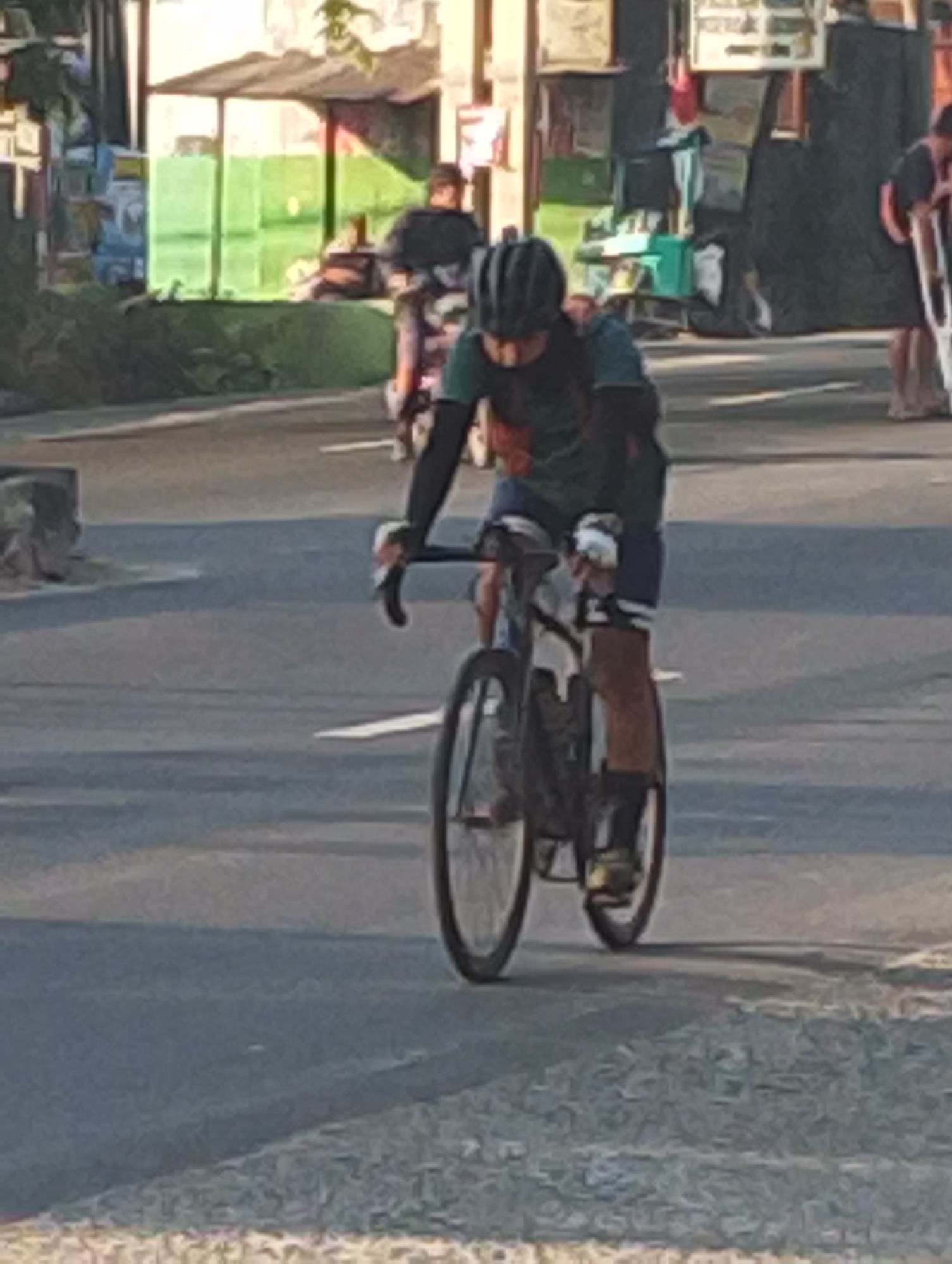 A cyclist wearing a helmet and athletic clothing rides a bicycle on a paved road. The cyclist appears to be wearing arm and knee pads. The background shows buildings and trees, suggesting an urban or semi-urban environment. There are other people visible in the background, some on bicycles and some standing. The lighting suggests it is daytime, possibly late afternoon due to the long shadows. The scene is consistent with a street in Gempol, Indonesia, which is known for its busy roads and commercial areas.