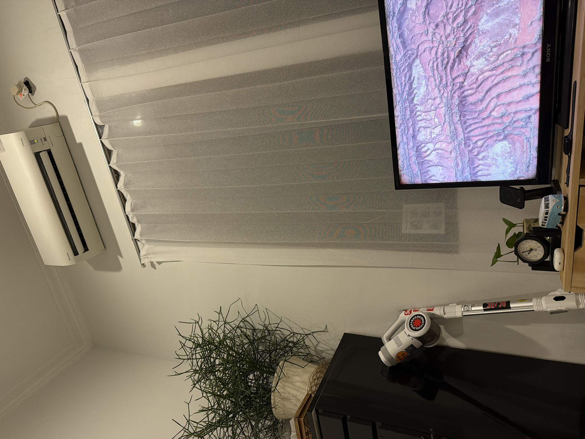 The image shows an indoor scene, likely a living room or bedroom, in East Jakarta, Indonesia. The focal point is a black Sony television displaying a satellite image of a desert landscape, identified as "Desert, Hardap, Namibia" from Google Earth. To the left of the television is a bedside table with a small plant, a clock, a toy Volkswagen van, and an Apple AirPod case. Leaning against a white wall next to the television is a white cordless vacuum cleaner, adorned with several stickers. Further left, a dark dresser is partially visible, topped with a potted plant with thin, branching stems. Above the dresser and plant, a white Daikin air conditioning unit is mounted on the wall. Translucent white curtains hang over a window to the right of the air conditioner. The lighting suggests it is nighttime or a dimly lit room.
