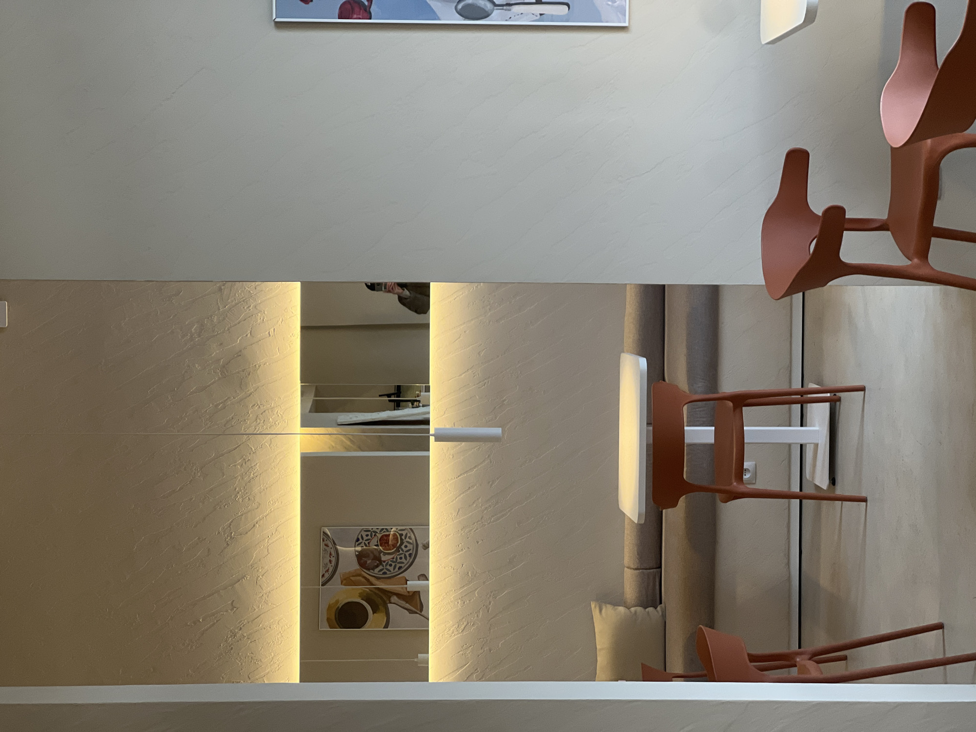 This media file captures a modern interior space, likely a dining or living area, characterized by clean lines and textured surfaces. The image is oriented sideways, presenting a vertical view of the room.
On the left side of the frame, a textured, light beige wall with subtle patterns is illuminated by a hidden vertical strip light running along its edge. A tall, narrow mirror or reflective panel is positioned to the left of this light, reflecting elements of the room. A person holding a camera or smartphone is visible in the reflection, indicating they are taking the photograph. Below this reflection, a framed picture or decorative tile depicting a meal (possibly a plate with food and a coffee cup) is reflected on a shelf. Above the textured wall, a plain light grey wall is visible, with the top portion of a framed artwork showing a red object and a metallic spoon.
The right side of the image features two modern, reddish-brown (terracotta-colored) chairs with organic, branching frames, stacked vertically against what appears to be a wall or a structural element. Another similar chair lies horizontally on a lower surface, its legs extended. Behind these chairs, a light-colored, upholstered cushion or panel is visible. A white, rectangular wall-mounted light fixture is positioned on the upper right.
The overall aesthetic is contemporary and minimalist, with a neutral color palette of light greys, beiges, and the reddish-brown accent from the chairs. The warm glow from the hidden strip lighting suggests an indoor setting, likely in the evening or a controlled lighting environment. The activity depicted is primarily a person documenting the interior space. The location context provided is Khimki, Russia.