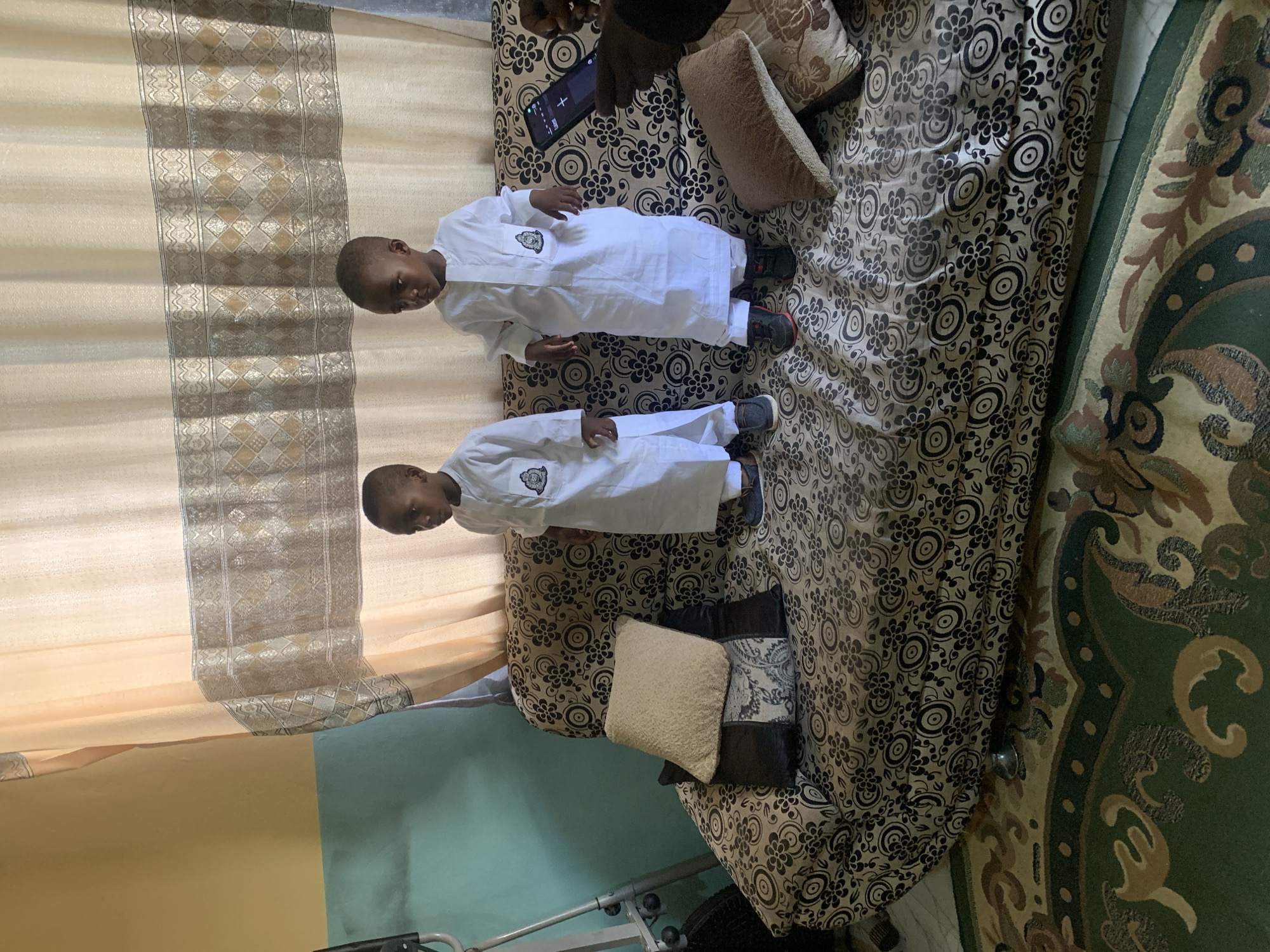 Two boys in white garments lie on a patterned sofa. An arm with a phone is visible at the top. The sofa sits on a patterned rug. A wall is visible in the background. The location is Gura Top, Nigeria.