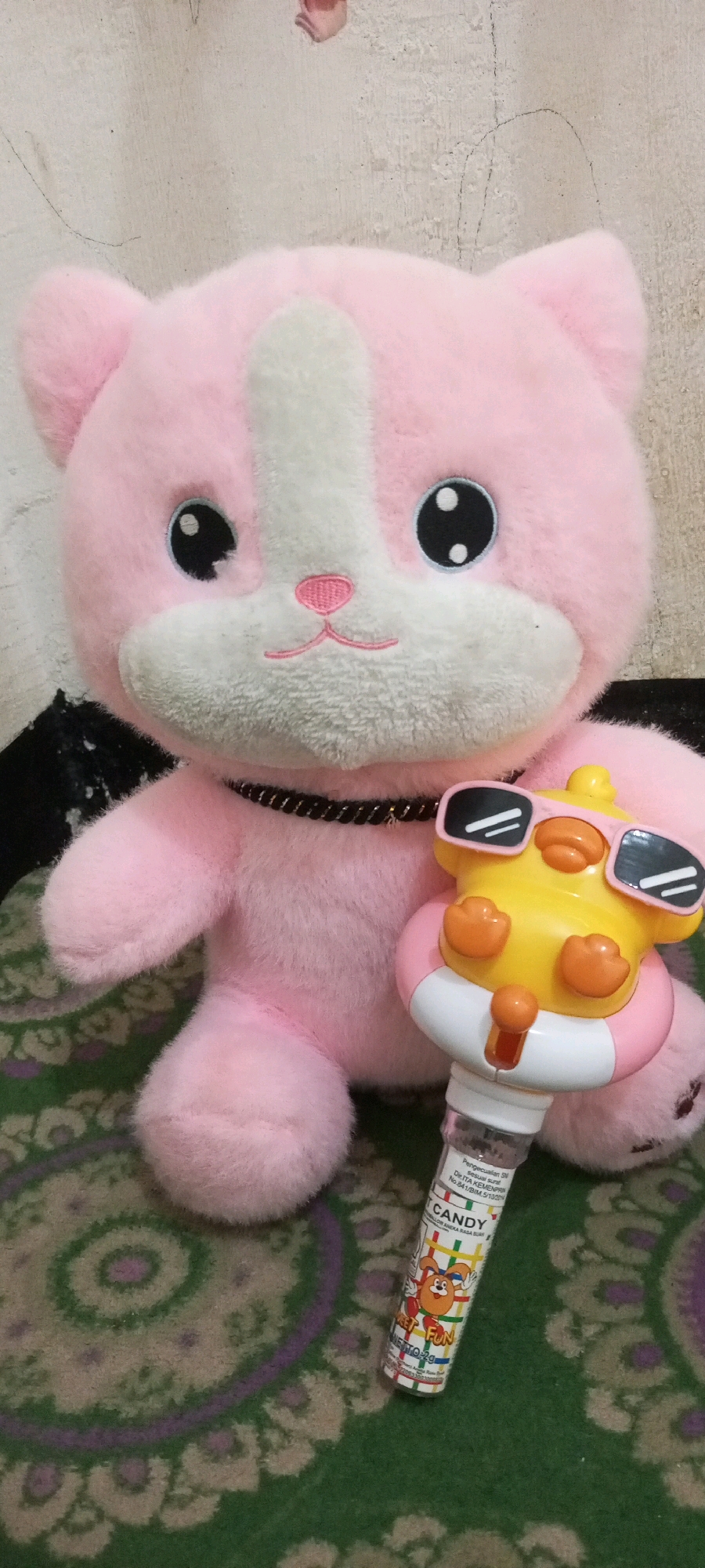 A pink stuffed cat is positioned in the center of the frame, seated on a green rug with a circular, floral pattern. The cat is adorned with a black, braided collar. In front of the cat and to its right, a yellow toy duck with sunglasses is held by a clear plastic tube containing small colorful candies. The duck toy is wearing pink and white sunglasses and has orange accents. The plastic tube has text and images on it, including the word "CANDY" and a cartoon character. The background is a textured, off-white wall. The scene appears to be indoors, possibly in a home environment. The image does not depict any specific location within the city of Sendawar, Indonesia, or any activity that would suggest its location.