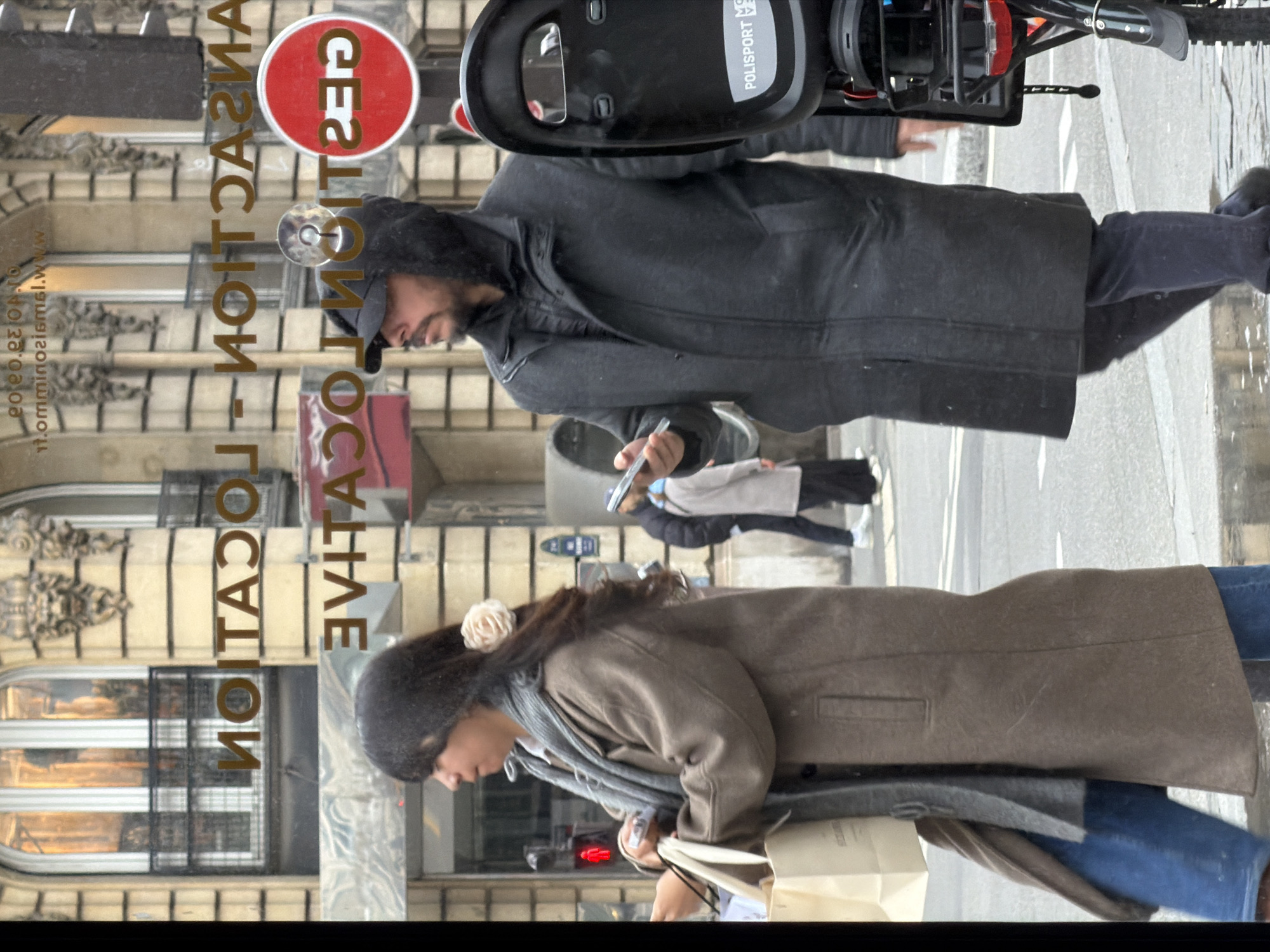 A street scene in Paris, France, depicts two individuals in the foreground, with others in the background. The person on the left is wearing a dark knit hat, a light-brown coat, a gray scarf, jeans, and is carrying shopping bags. The person on the right, situated higher in the frame, is wearing a hooded black coat and jeans, while holding a smartphone. The scene appears to be captured through a store window displaying the reversed text "LOCATION" and a "NO ENTRY" sign. A black device, possibly part of a vehicle, is positioned at the top. The background shows other pedestrians walking on the street.