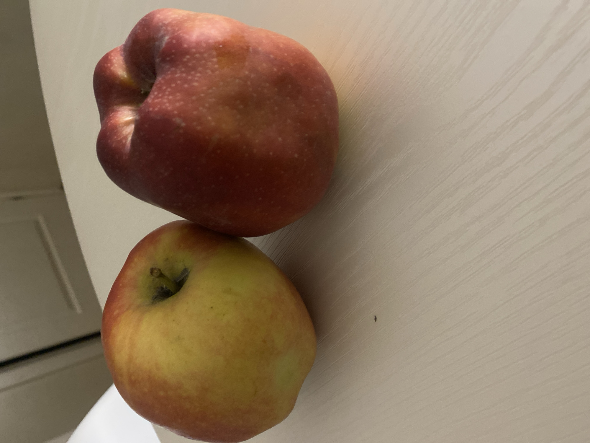 Two apples sit on a light-colored, textured surface. The apple on the left is primarily yellow with red blush and has its stem. The apple on the right is predominantly red with some yellow undertones and appears slightly misshapen. A small, dark speck is visible on the surface between the apples. The background is out of focus and shows a hint of cabinetry or wall.