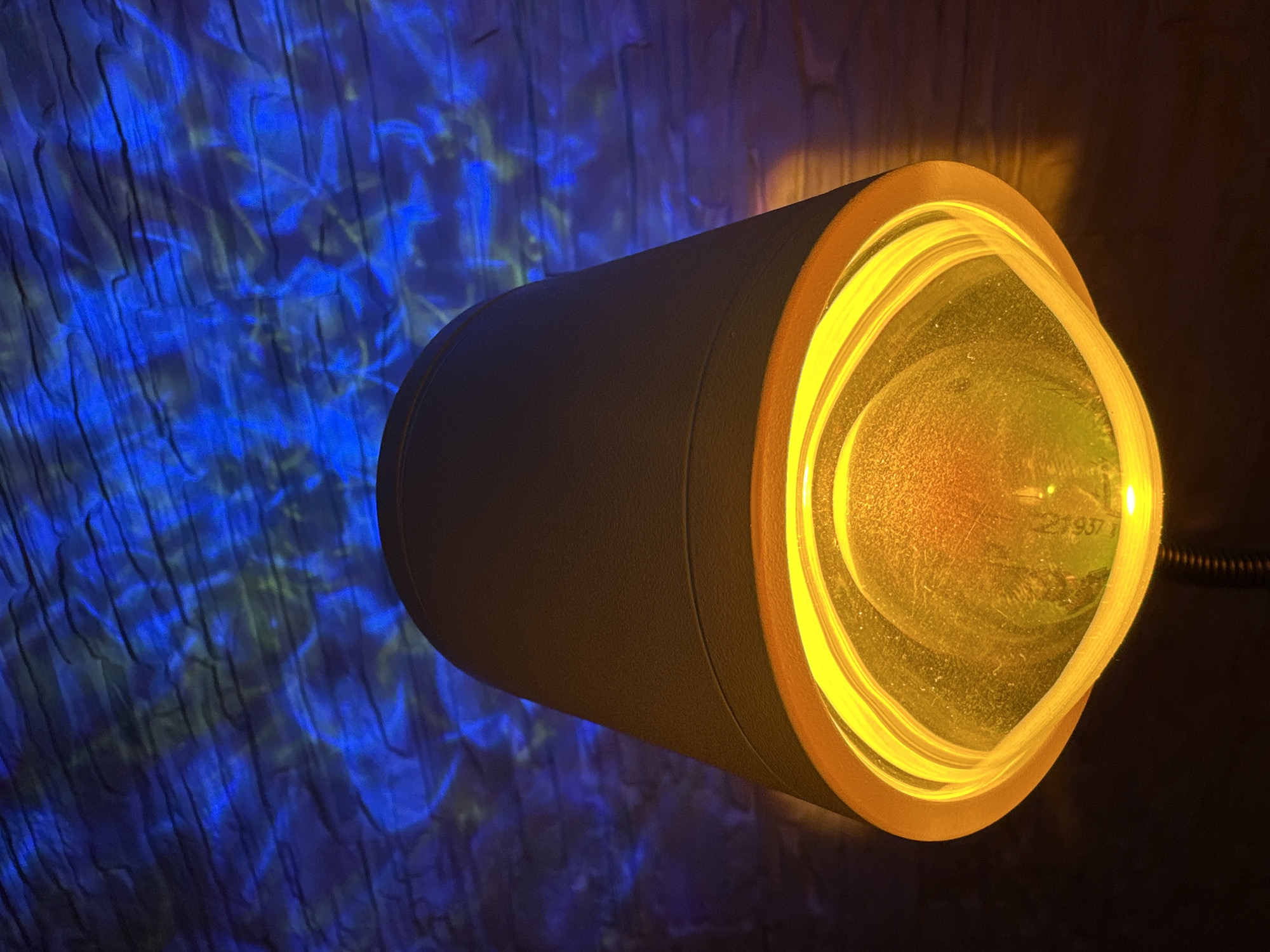 A cylinder-shaped, tan-colored light fixture with a bright yellow lens is mounted on a dark wall. The lens of the light fixture projects swirling patterns of blue and green light onto an adjacent wall. The location is unknown.