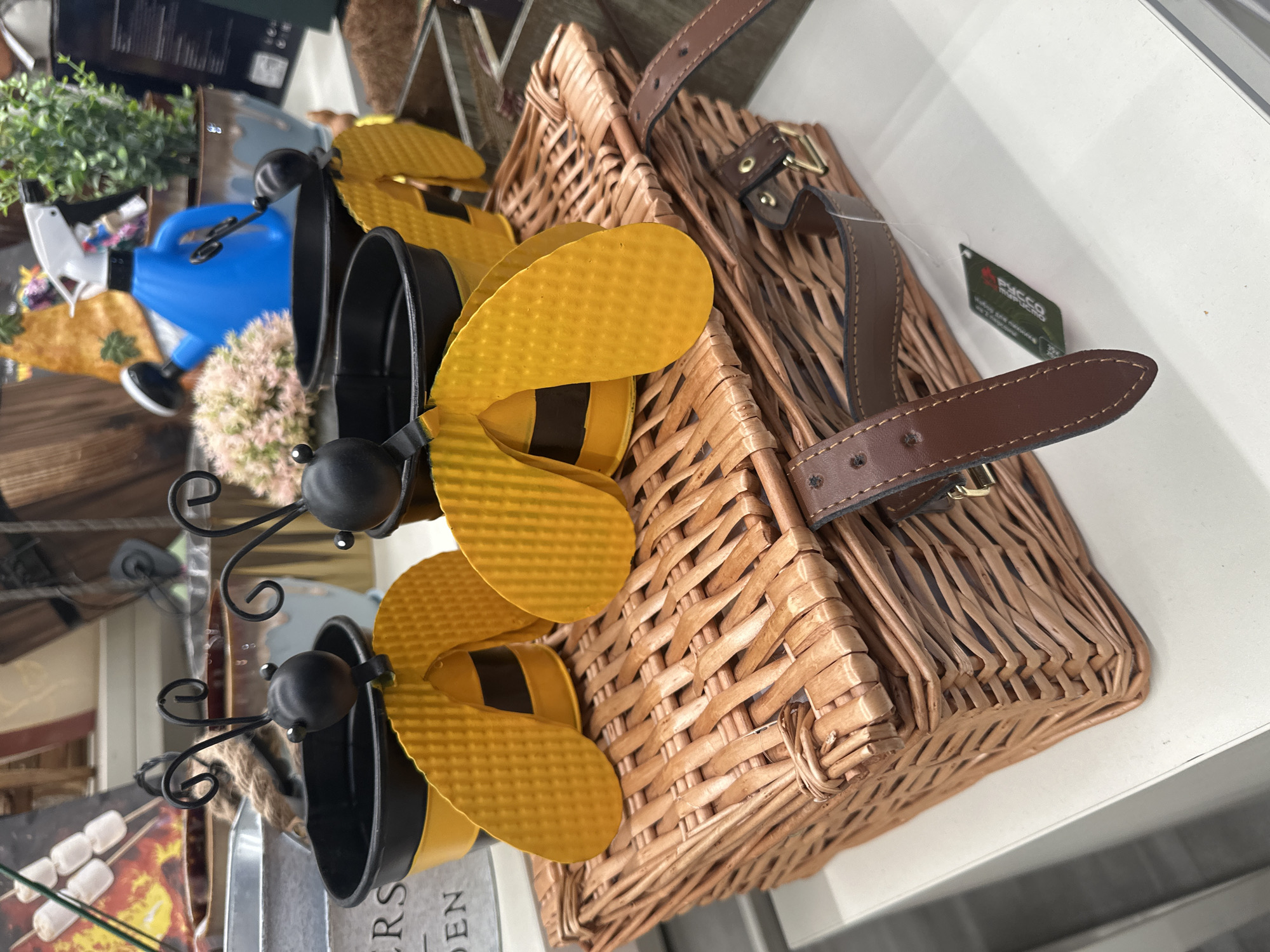 In an indoor setting, possibly a store, a wicker picnic basket sits on a white surface. The basket is adorned with decorative bee-shaped planters, each featuring yellow wings and black stripes. These planters are positioned on top of the basket. In the background, various gardening-related items are visible, including a blue watering can with a white nozzle, a plant in a decorative pot, and what appears to be a display of marshmallows on skewers. The scene is well-lit, suggesting it's a product display.