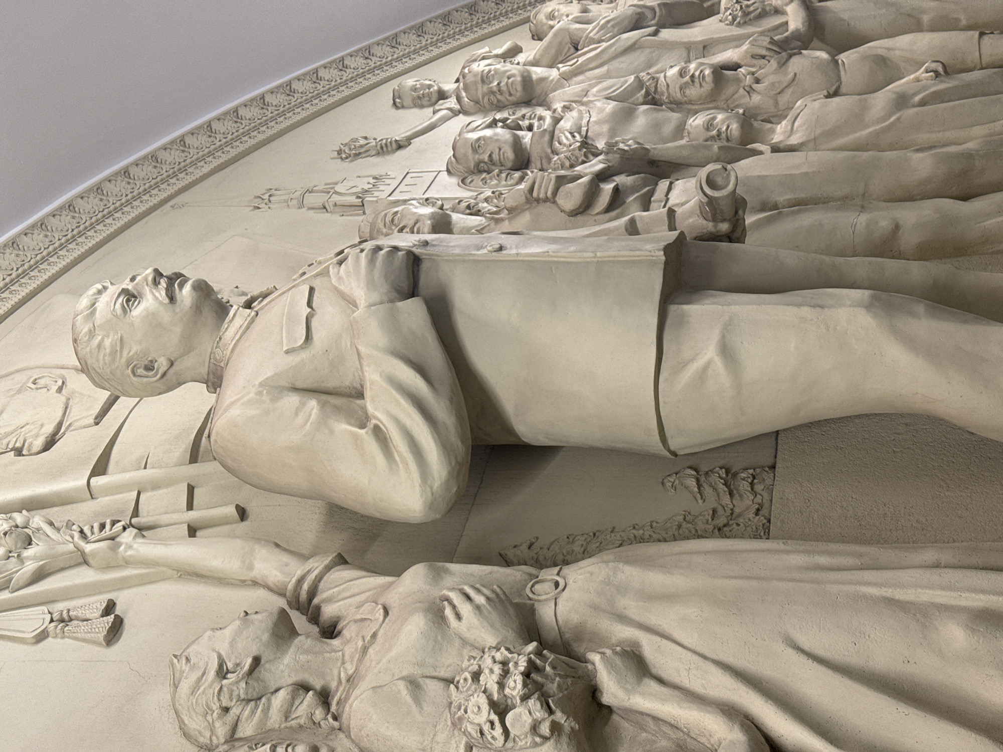 The image captures a detailed bas-relief depicting fallen heroes, located in the Moscow Metro, Russia. Several figures are shown lying down, seemingly lifeless, with peaceful expressions. The most prominent figure is a man in a suit, his head turned upward. Below him lies a woman in period clothing. Above the man in the suit are figures, including a young girl, all positioned as if in repose. A decorative border is seen at the top of the relief.