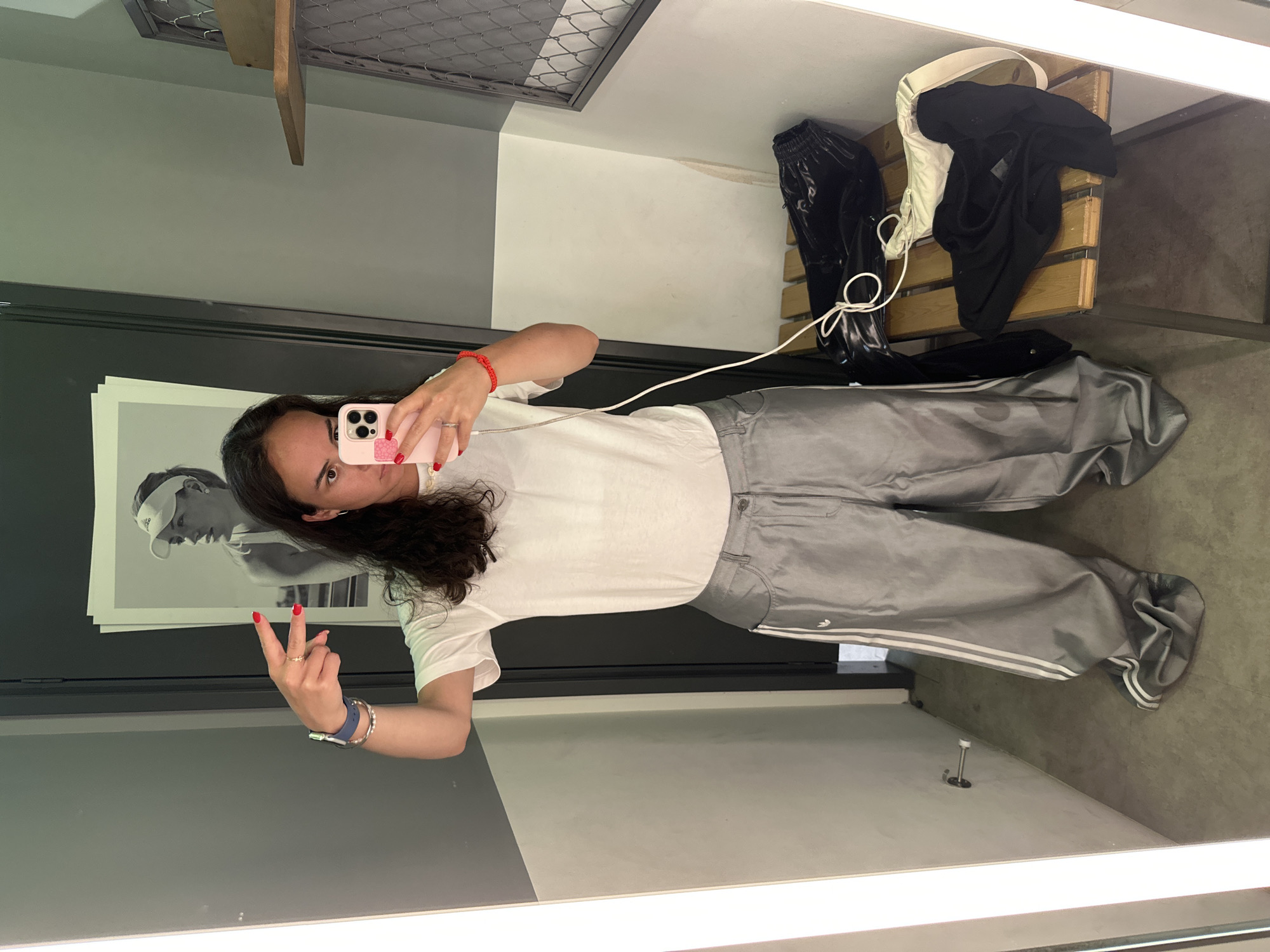 A person is taking a picture in a fitting room. The person is wearing a white t-shirt and grey Adidas track pants and holding a pink smartphone. Their left hand is extended, forming a "peace" sign. The fitting room has grey and white walls. A picture of a tennis player is on the wall. A wooden bench with clothing is in the background. The location is possibly in a store in Pattaya City, Thailand.