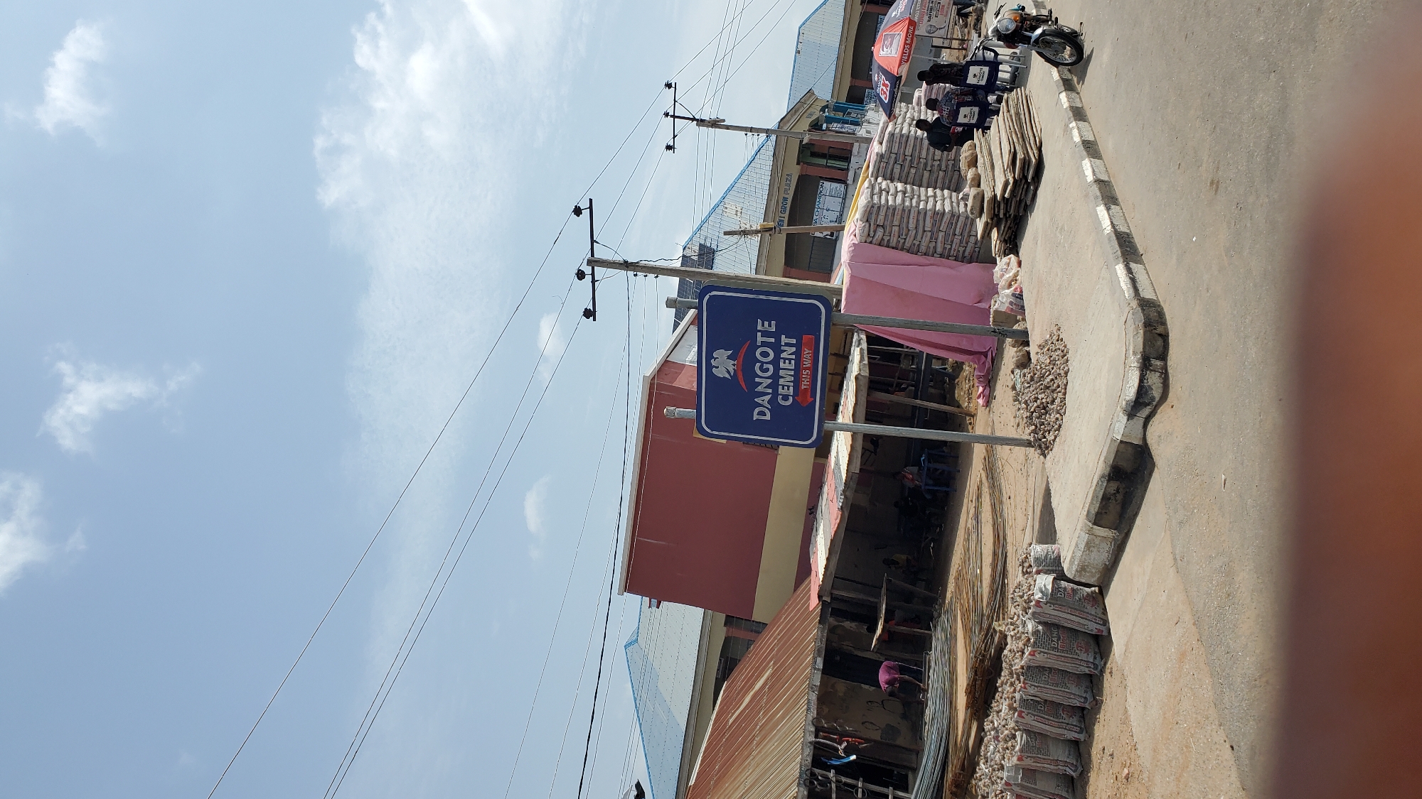 The image depicts a street scene in Jalingo, Nigeria, likely an outdoor market or commercial area. In the foreground, a sidewalk with a concrete curb is visible, with some debris scattered on it. A prominent blue sign with white text and an emblem indicates "DANGOTE CEMENT THIS WAY," suggesting a directional marker for the cement company's premises or distribution point.
To the left of the sign, there are makeshift stalls with corrugated metal roofs, housing stacks of rebar and bags of what appears to be cement. Several people are present, some standing and engaged in activities, while others are seated under the shelters. A large pink tarp is draped over some of the materials.
Further back, buildings with blue roofs are visible, one with a red facade. In the background, there are more structures, including one with signage that reads "CEMENT GROW PLAZA." Power lines crisscross the sky. To the far right, a motorcycle is parked on the roadside, and a few individuals are seated near umbrellas, one of which has visible branding. The sky is mostly clear with scattered white clouds. The overall impression is a bustling commercial environment with an emphasis on building materials.