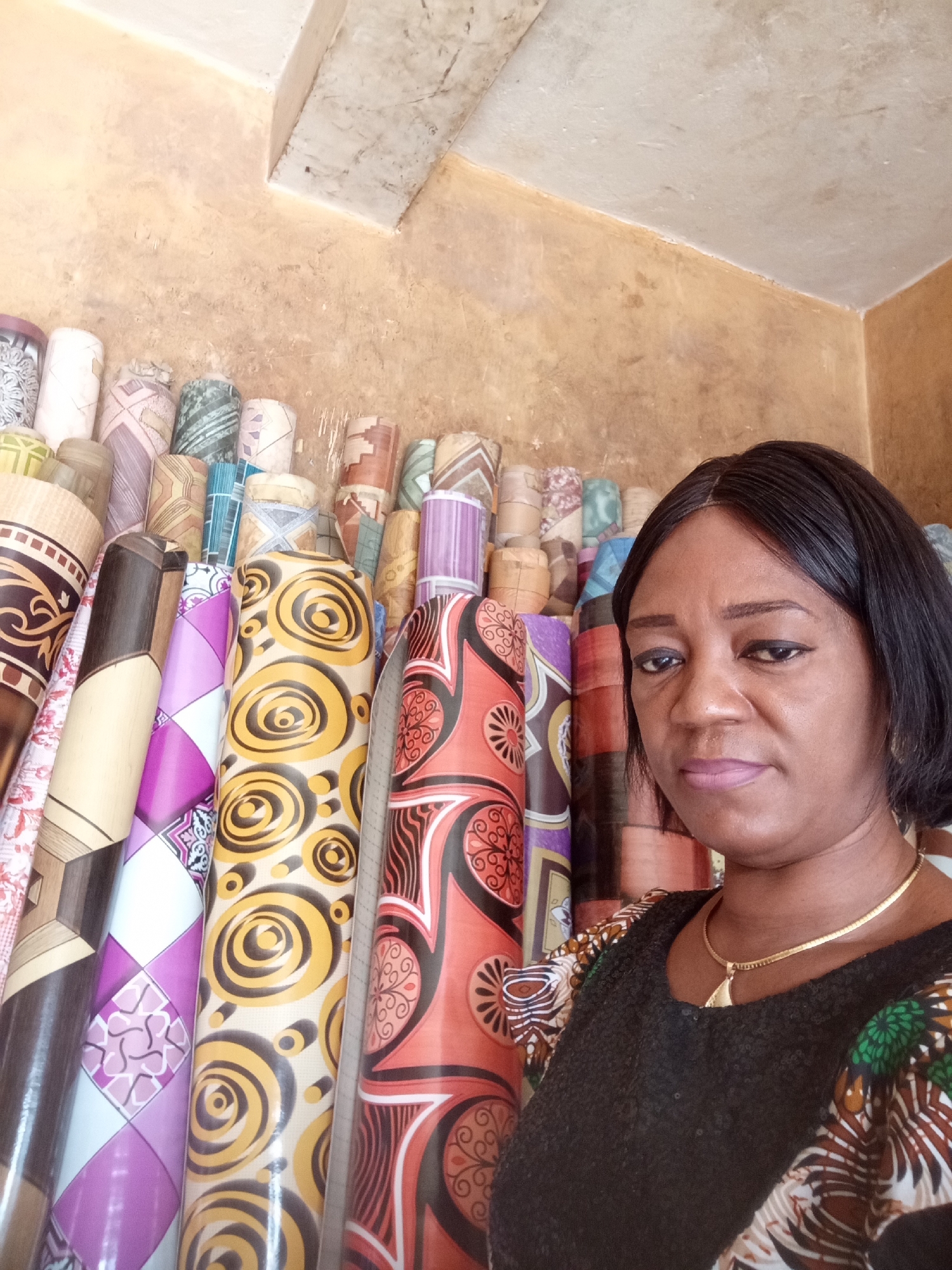 A woman stands prominently on the right side of the frame, looking directly at the camera with a neutral to slightly serious expression. She has short, dark hair and is wearing a dark top with colorful, patterned sleeves and a gold chain necklace with a small pendant. Her lips are a light pink.
Behind her, and filling most of the background, are numerous rolls of patterned floor coverings, likely linoleum or vinyl. These rolls are stacked vertically and horizontally, creating a vibrant and busy backdrop. The patterns on the rolls vary widely, featuring geometric designs, swirling motifs, and abstract shapes in a diverse palette of colors including yellow, black, red, orange, purple, white, brown, and green.
The setting appears to be an indoor shop or market stall in Maiburiji, Nigeria, specializing in home floor coverings. The walls are a warm, earthy tone (light brown or tan), and the ceiling is a lighter, possibly concrete or painted surface. The lighting suggests it is daytime. The image functions as a portrait of the woman within her business environment, highlighting her alongside the products she sells. No visible text is present in the image.