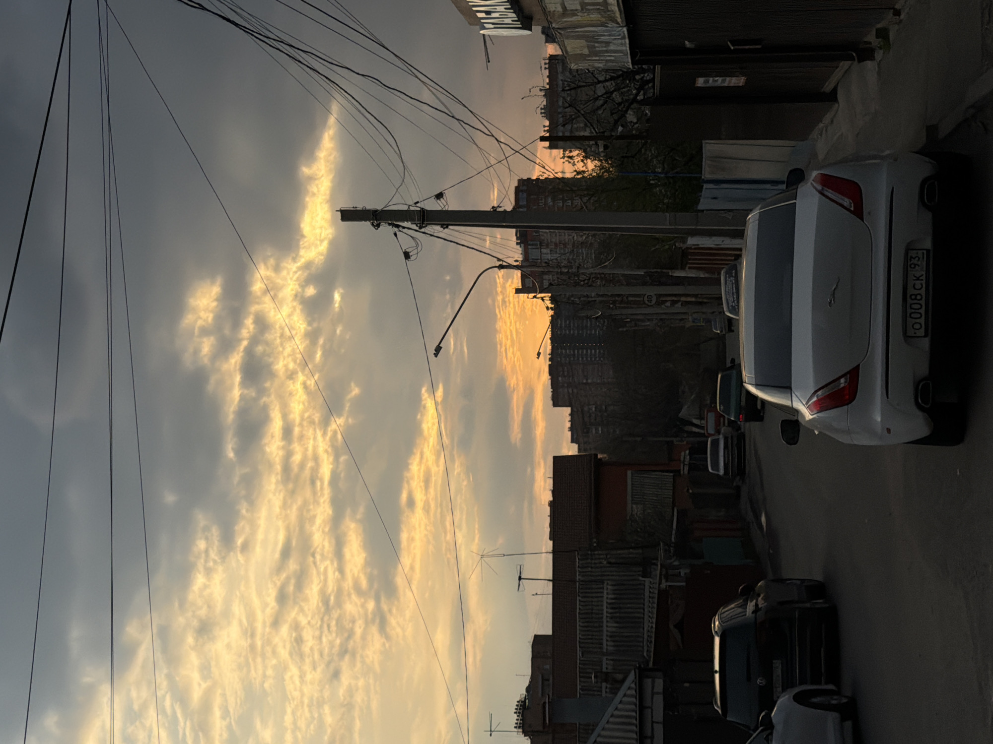 The image depicts a street in Rostov-on-Don, Russia, at sunset. The sky is a mix of gray and orange, with elongated streaks of orange clouds. A utility pole with various wires stands in the foreground. Several cars are parked along the side of the road. A white Maserati with the license plate "O 008 CK 93" is prominently featured. Buildings of varying heights line the street.