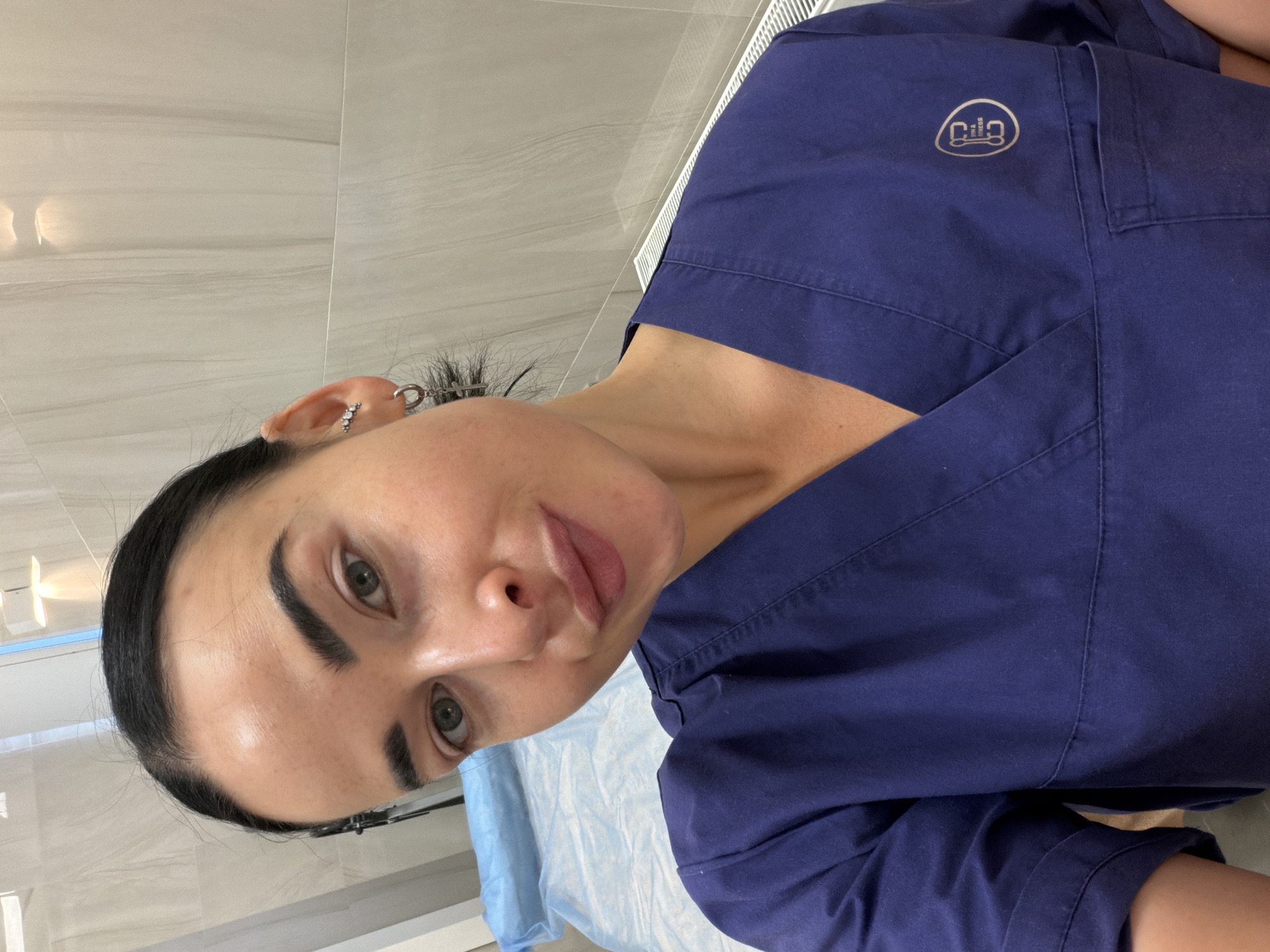 A woman with dark hair pulled back and wearing a dark blue scrub top with a circular logo on the upper right chest area is looking at the camera. She has fair skin, defined eyebrows, and multiple piercings in her left ear. The background appears to be a clinical setting with light-colored, textured walls. A light blue sheet is visible on what appears to be an examination table. The location in the city of Saint Petersburg, Russia, is unknown.