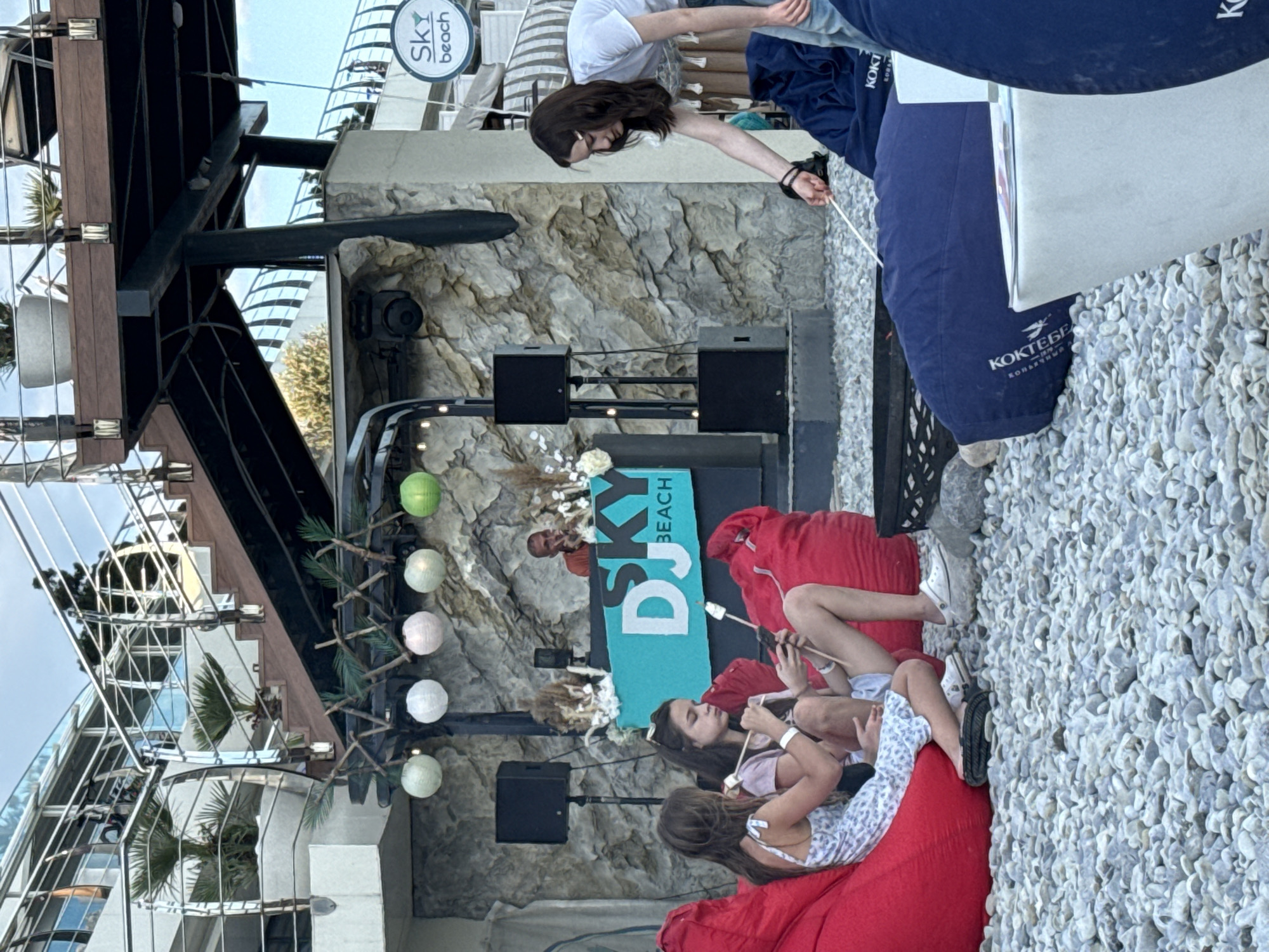 The image depicts a group of young women gathered on a beach, sitting on large red beanbag chairs. The scene is set at a beach club, indicated by a prominent "DJ SKY BEACH" sign. The women appear to be enjoying a relaxed social gathering, with some holding skewers and one woman looking at her phone. The lighting suggests it's daytime, possibly late afternoon, with a clear sky.
Notable details include the rocky beach surface, a stage with speakers and a DJ booth, and decorative round lights hanging from a structure. The background shows a multi-story building with glass railings, reflecting the sky and greenery. There is also a large blue tarp or blanket with visible text, including "KOKTEBE" which is a location in Crimea, suggesting this is the general area. One woman is holding a white string, possibly interacting with a prop or a game. The overall mood appears casual and celebratory.
