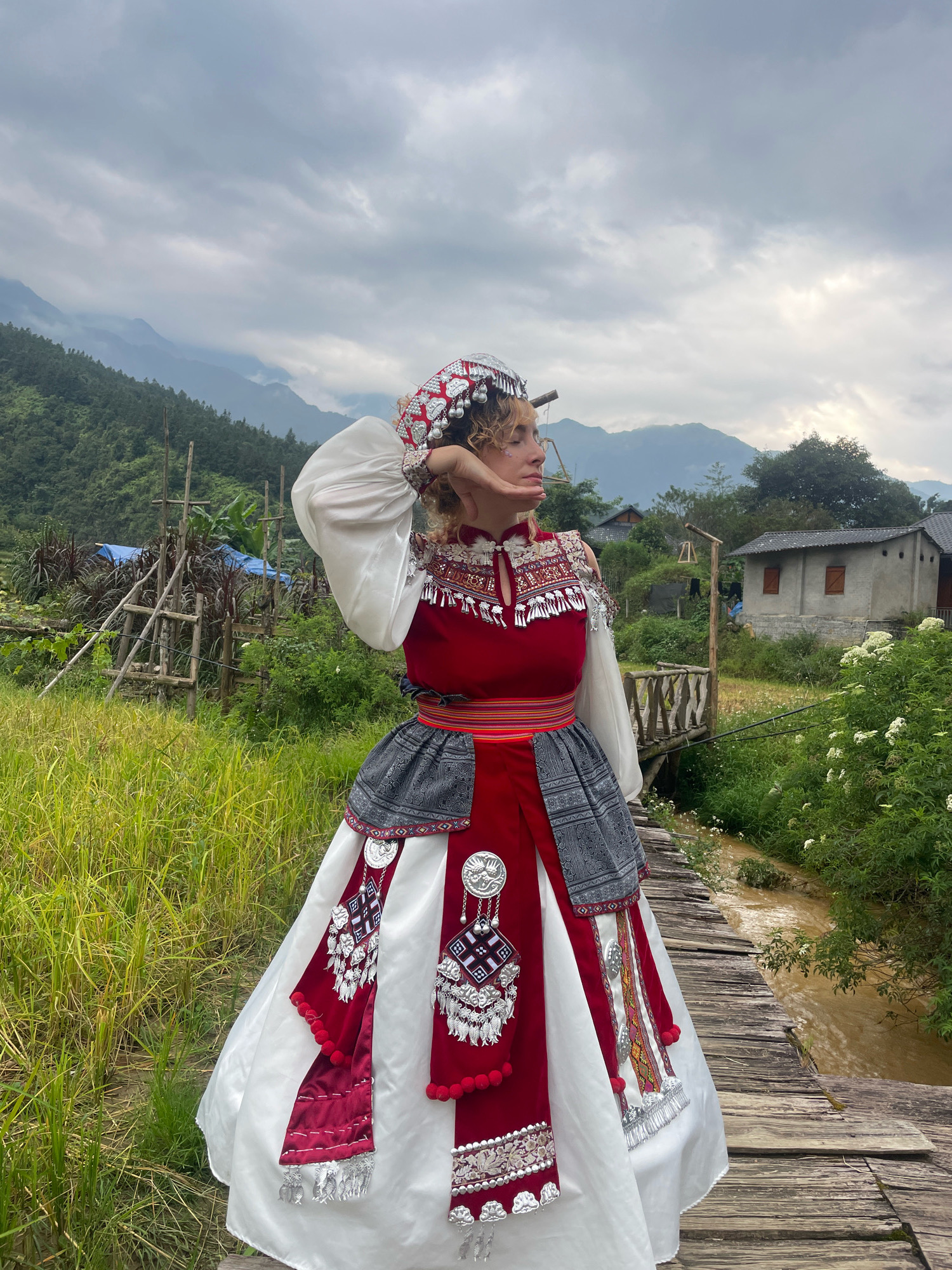 A woman stands on a wooden bridge in Cat Cat Village, Vietnam, wearing a red and white traditional outfit adorned with intricate silver embellishments and a decorative headdress. The background features lush green rice paddies, a small stream, a rustic wooden structure with blue tarp, and houses nestled amidst rolling hills under a cloudy sky. The woman's pose and gaze suggest she is looking away from the camera, possibly towards the landscape. The overall scene depicts a rural, ethnic setting.