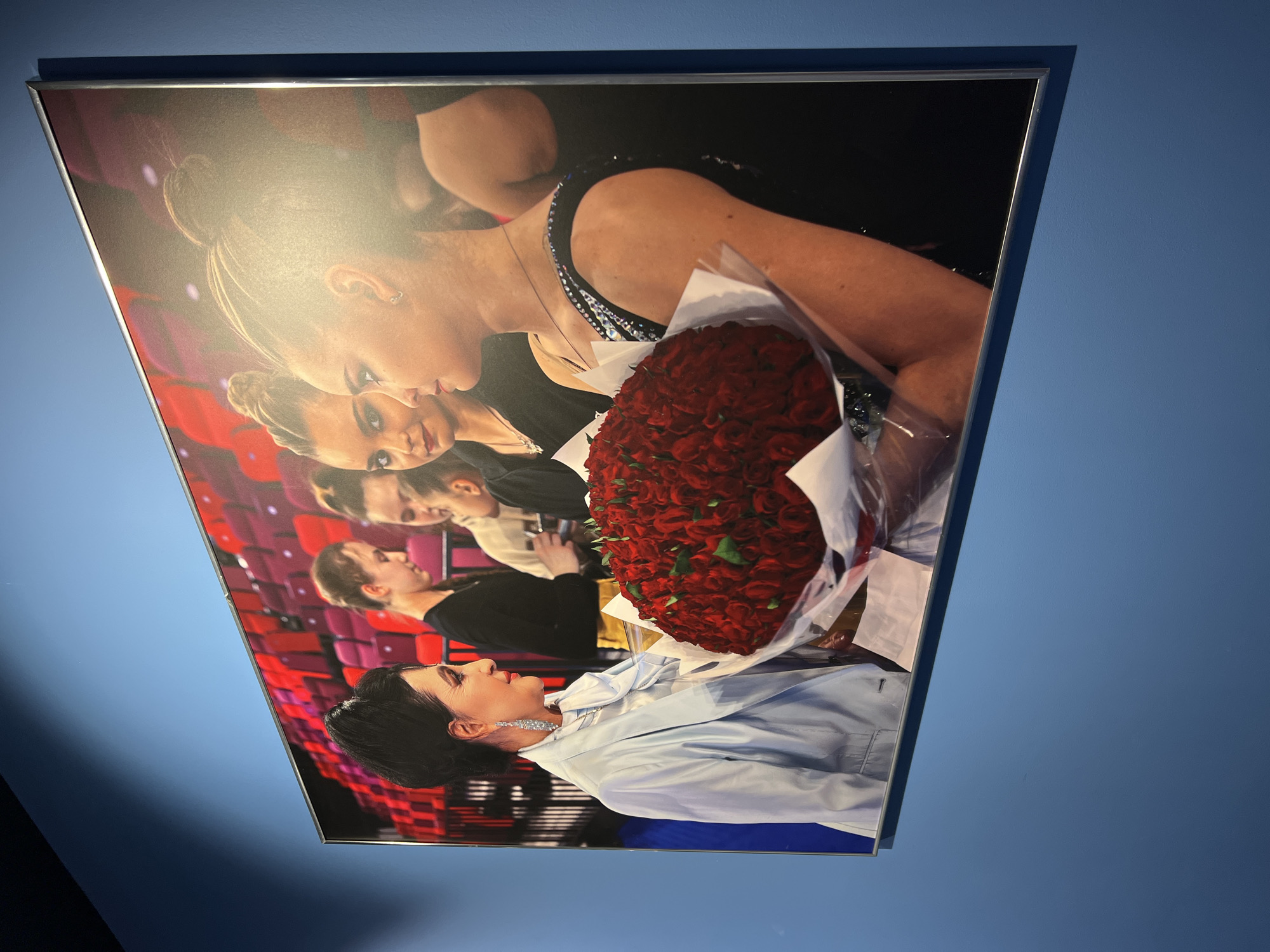 A framed photograph hangs on a blue wall, depicting a scene with several individuals and a large bouquet of red roses. In the foreground, on the left, a woman with dark, upswept hair and prominent dangling earrings wears a light blue blazer over a white collared shirt and a scarf. She is looking to her right, her head slightly tilted. In front of her, a substantial bouquet of red roses, wrapped in clear plastic and white paper, is held by two young women. The woman on the right, facing left, has her hair pulled back and is wearing a dark, sleeveless dress with embellishments on the shoulder strap. Her arm is visible holding the bouquet. To her left, another young woman with her hair also pulled back is partially visible, looking towards the bouquet. Behind them, other figures and red seats in what appears to be an auditorium are visible. This photograph is located in Moscow, Russia.