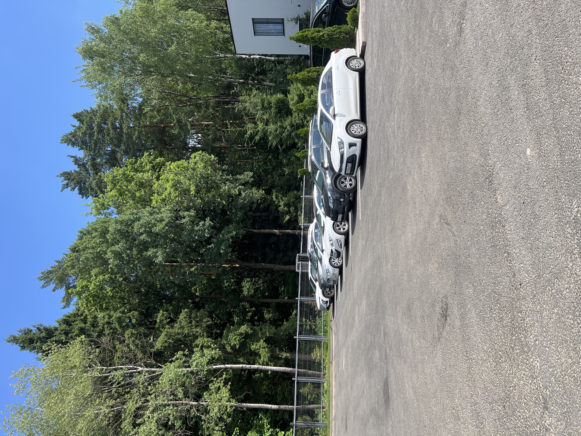 A white sedan, a dark SUV, a silver sedan, and two other silver sedans are parked in a lot. The lot is paved with asphalt and is bordered by a metal fence and a forest. A modern white building with a dark-trimmed window is visible to the right of the parking lot. The sky is clear and blue. The location is Минзаг, Russia.