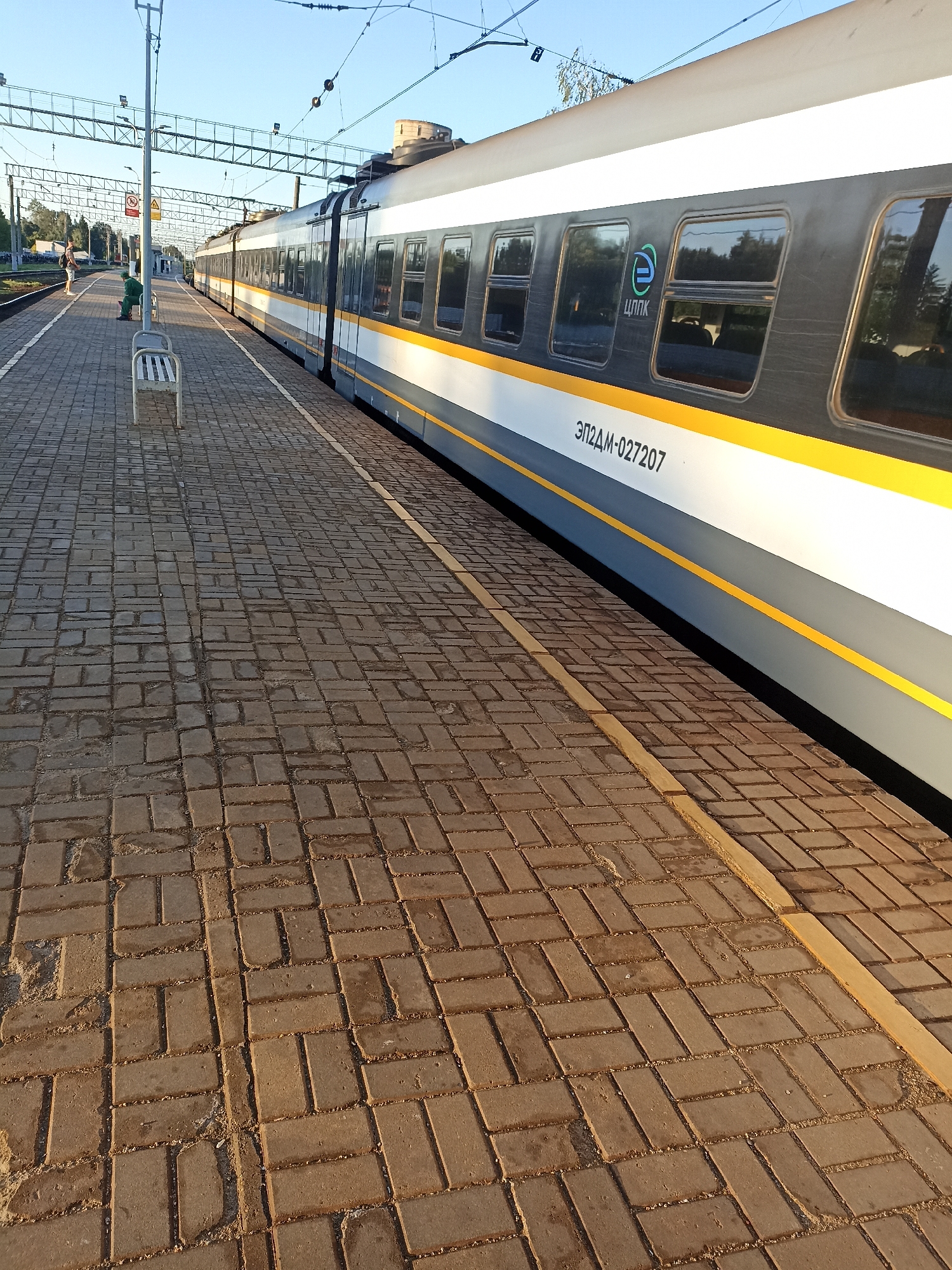 An outdoor, eye-level, wide shot captures a train at a station in Chekhov, Russia. The train, running parallel to the foreground, is dark gray with a white and yellow stripe along its side. Several windows are visible on the train, reflecting the surrounding environment. A logo with the letters "ЦППК" and a stylized "e" is present on the train's exterior. Below the windows, the text "ЭП2ДМ-027207" is printed. The platform is paved with rectangular bricks laid in a herringbone pattern. Two benches are visible on the platform, with one person sitting on one of them and another person standing nearby. Overhead, electrical wires and metal structures for the train's power supply are visible against a clear blue sky. The sun casts long shadows across the platform.