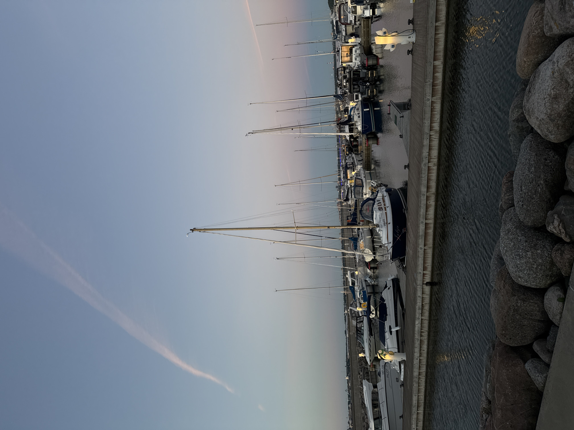 This is an outdoor, horizontal, medium-wide shot of a marina at dusk in Tallinn, Estonia. The sky is a soft gradient of pale blue to a very faint pinkish-orange near the horizon, with a prominent, thin contrail stretching diagonally across the upper left quadrant of the sky.
In the foreground, large, rounded rocks are visible along the edge of the water. The water itself is dark and rippling, reflecting the dim light and lights from the marina. A wooden pier extends into the water from the right, with several mooring posts and a tall, modern-looking light fixture emitting a warm glow.
The marina is filled with a variety of docked boats, mostly sailboats and some smaller motorboats. Many of the boats are white or dark blue, with some covered with tarps. The masts of the sailboats create a dense forest of vertical lines against the sky. Lights are visible on some of the boats and along the pier, casting reflections on the water. In the distance, the skyline of Tallinn is faintly visible, with buildings and structures silhouetted against the horizon. The overall atmosphere is tranquil and serene, indicative of a quiet evening in a port.