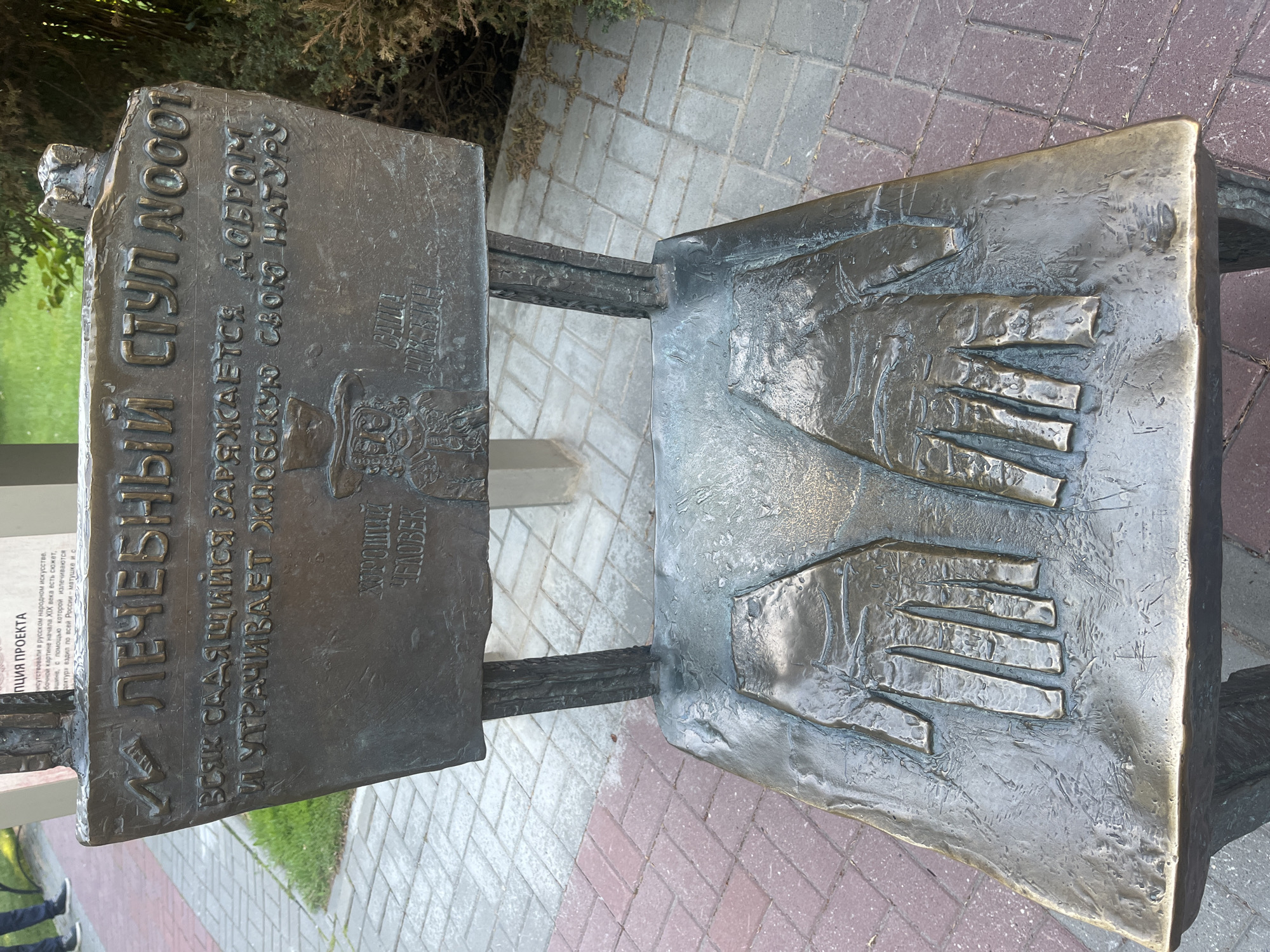 This media file features a bronze sculpture of a chair with two prominent hand imprints on its seat, one positioned above the other. The sculpture is situated outdoors on a paved area with a mix of gray and reddish-brown interlocking bricks. To the left of the chair, a metal plaque is attached, also made of bronze, bearing text in Russian. The plaque is mounted on a vertical support that is part of the chair's structure.
The text on the plaque reads: "ЛЕЧЕБНЫЙ СТУЛ А0001 ВСЯК САДЯЩИЙСЯ ЗАРЯЖАЕТСЯ ДОБРОМ И УТРАЧИВАЕТ ЖЛОБСКУЮ СВОЮ НАТУРУ ХОРОШИЙ ЧЕЛОВЕК" which translates to "Healing Chair A0001 Whoever sits down is charged with good and loses their spiteful nature. A good person." Below this main inscription, there is smaller text referring to a project and the connection of Russian folk art to 19th-century art. A faint image of a person appears on the plaque as well.
The background shows greenery, suggesting a park or garden setting, with some dry leaves on the ground near the base of the sculpture. The lighting indicates it is daytime, with no strong shadows suggesting a bright sun. The overall impression is of a public art installation designed to convey a message of positivity and well-being. The sculpture appears weathered, indicative of being exposed to the elements over time.