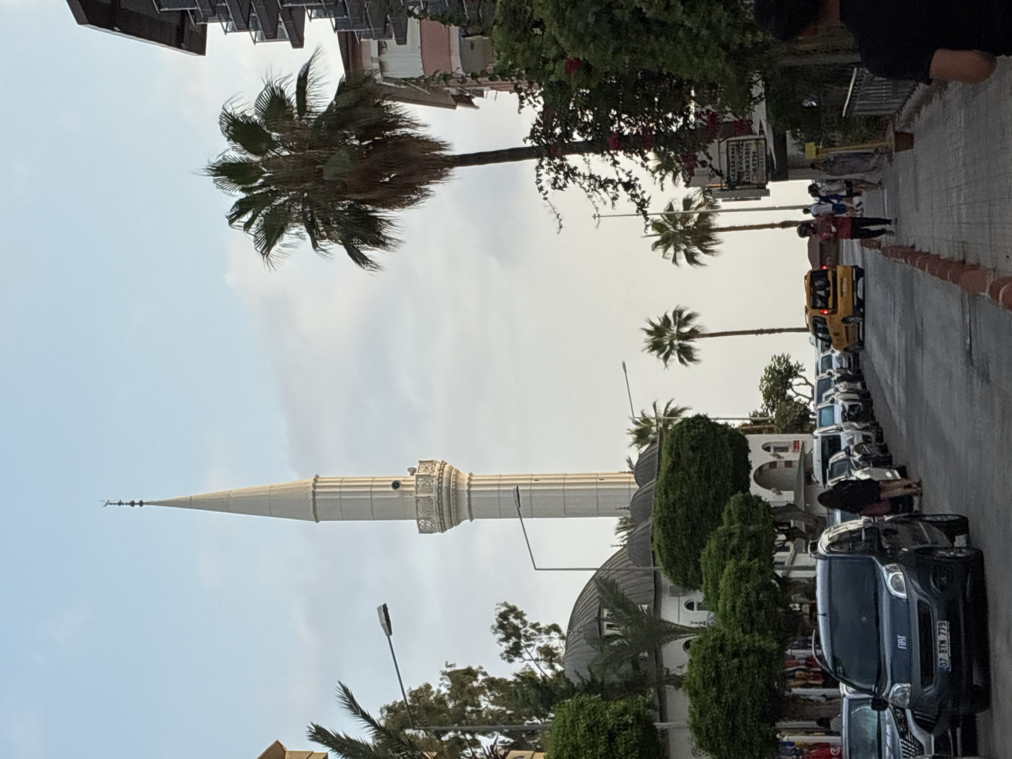 A street scene in Alanya, Turkey, features a prominent mosque minaret and dome on the left, with palm trees and buildings lining the right side of the road. Cars are parked along the street, and several people are walking on the sidewalk and crossing the road. The sky is overcast, and the overall lighting suggests it is either early morning or late afternoon. The mosque appears to be a significant landmark in the area, possibly located in a commercial or residential district, given the presence of shops and apartment buildings.