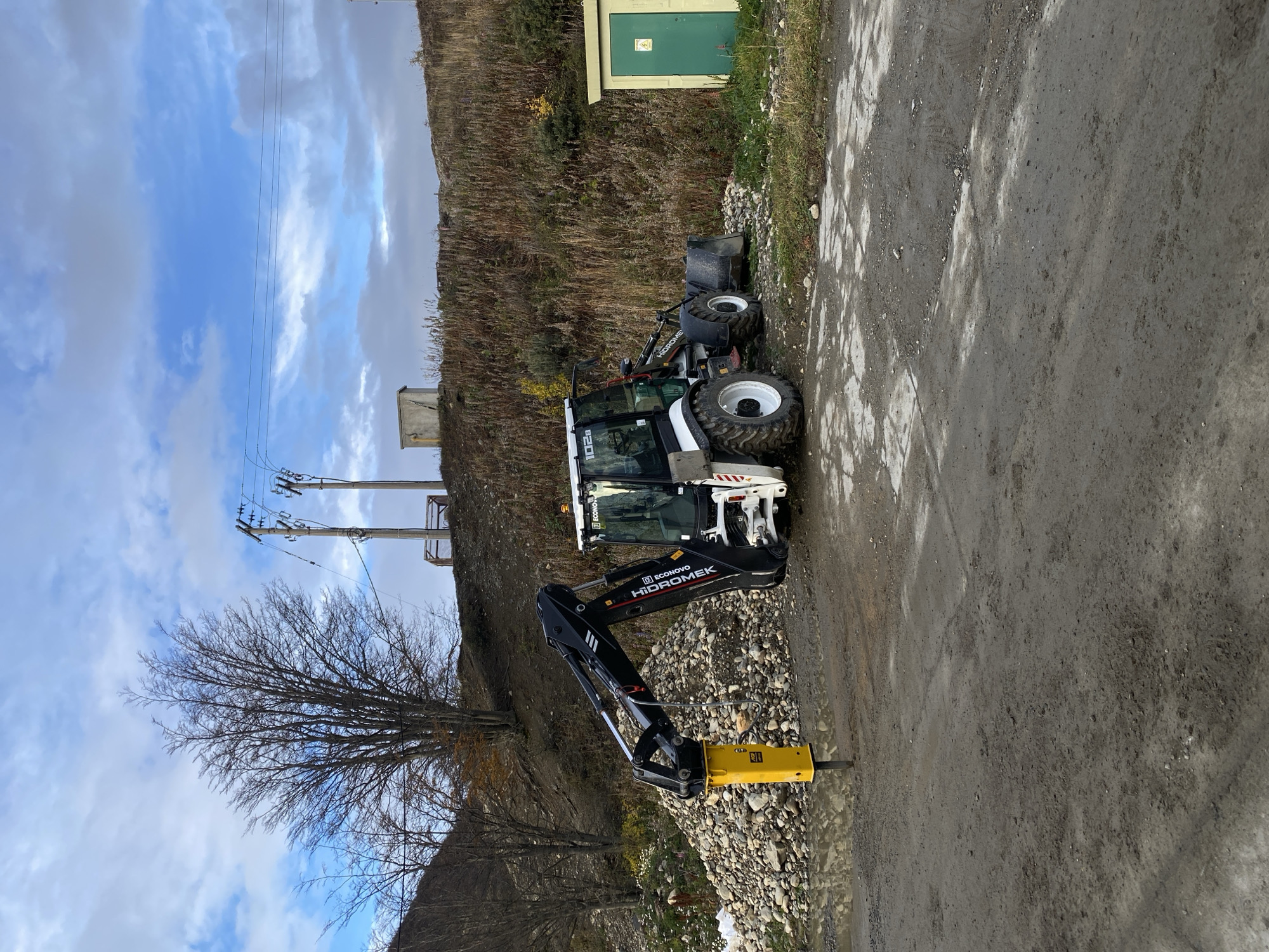 The scene depicts a dirt road or construction site near Ushuaia, Argentina. A white and black backhoe loader with the branding "HIDROMEK" is parked on a pile of rocks. The backhoe's hydraulic hammer attachment is extended towards the road surface. The surrounding environment includes a grassy hillside, a small green building, and a bare tree with visible power lines in the background. The sky is blue with scattered clouds.