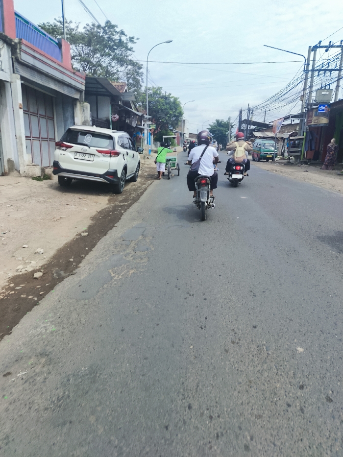 The scene is taken from a street in the city of Ciparay, Indonesia, located at latitude -7.024 and longitude 107.688. There are four people visible. Three are on motorcycles riding in the same direction. The fourth person is walking on the right side of the road. The motorcycle in the foreground is the closest to the camera. The person on the motorcycle is wearing a white t-shirt and a helmet. There is a white SUV parked on the right side of the road. The road is paved and has a slight curve to the right. There are houses on the left and right sides of the road. The buildings are low-rise and have a mix of residential and commercial uses. There are power lines and wires overhead. The sky is overcast and grey.