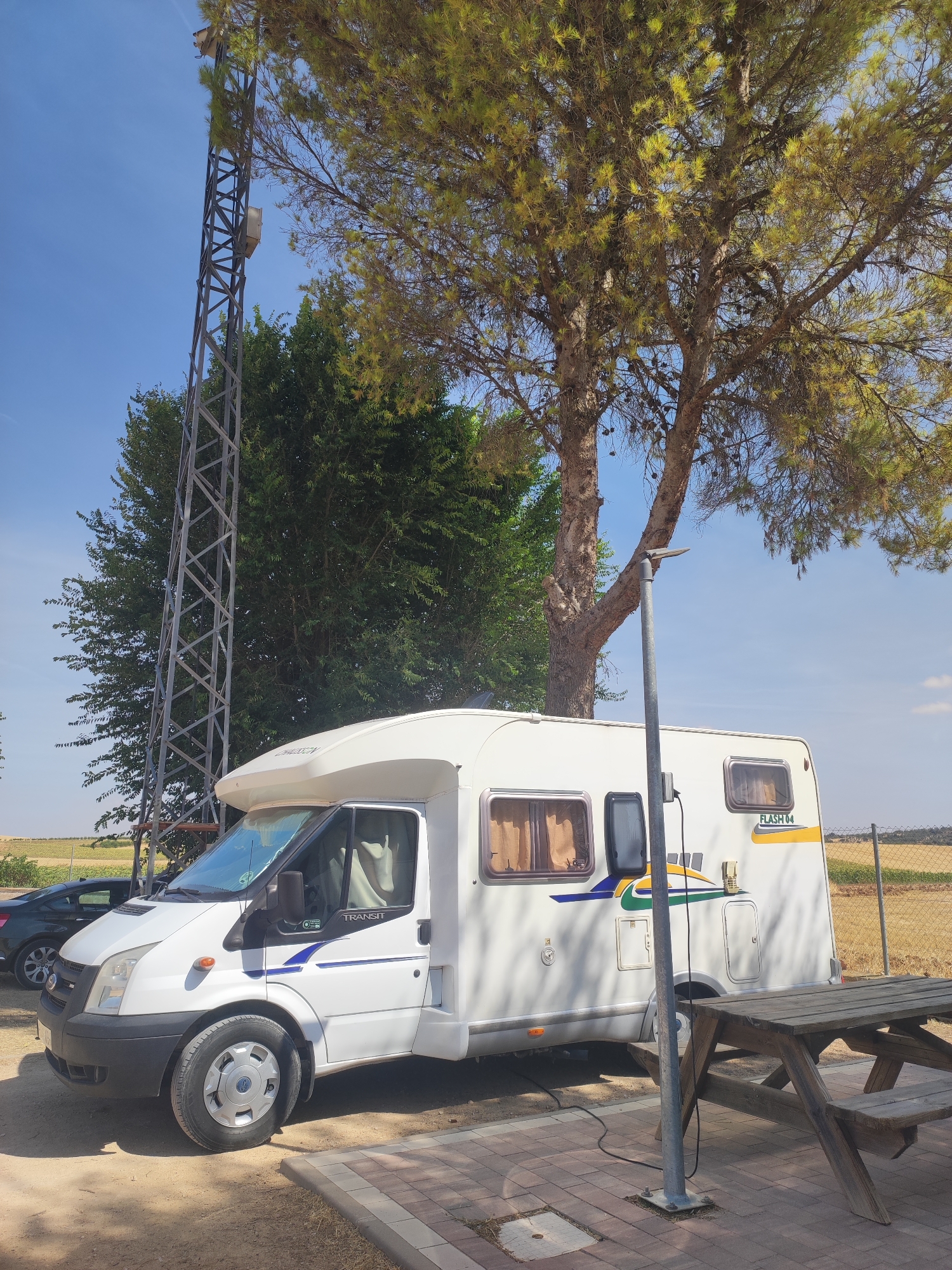 In Villarejo De Fuentes, Spain, a white camper van is parked on a dirt and paved area. The camper van has tinted windows and a decal that reads "FLASH 04". Next to the camper van is a wooden picnic table. Behind the camper van, a tall metal tower with lights on it rises into the blue sky. A large tree with green and yellow leaves partially shades the scene. In the background, there is a fenced-off field with dry grass. A dark-colored car is visible to the left of the camper van. The ground is a mix of dirt and paved tiles.