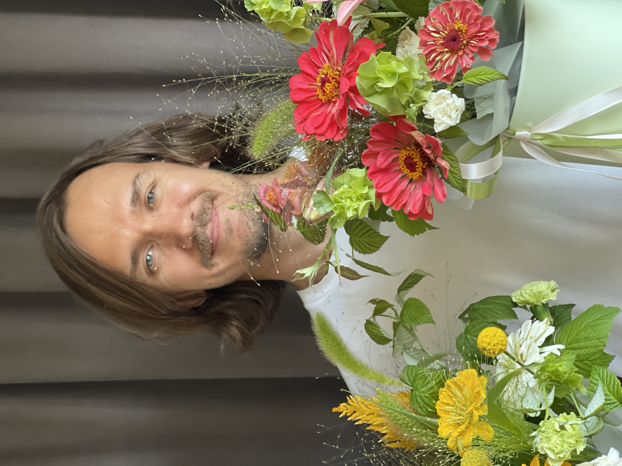 The image shows a man with medium-length brown hair and blue eyes, wearing a white t-shirt. He has a mustache and a small piercing on his nose. He is smiling and holding two bouquets of flowers. The bouquet on the left contains yellow and white flowers with greenery. The bouquet on the right, partially obscuring the man, features red zinnias, green hydrangeas, and other assorted flowers and greenery, presented in a light green box tied with ribbons. The background consists of a plain dark grey curtain. The setting appears to be indoors. The photograph was taken in Izhevsk, Russia.
