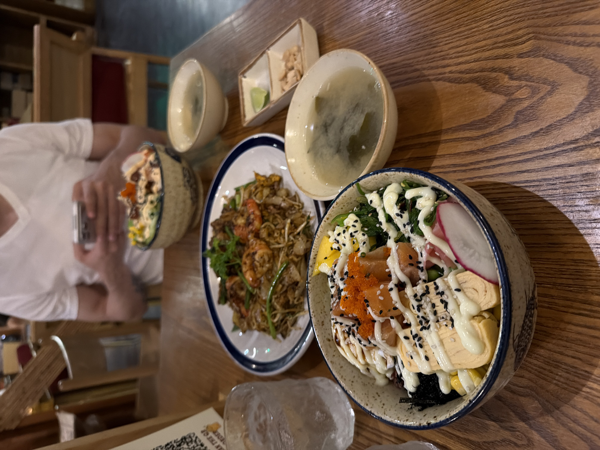 An eye-level, indoor shot on a wooden table shows a spread of Asian cuisine. From right to left, a patterned bowl of sushi or rice bowl is filled with salmon, seaweed, radishes, and a drizzle of white sauce. A white bowl filled with a dark, semi-transparent broth (possibly miso soup) is placed next to the bowl of sushi. Next to the bowl of broth is a small, divided dish containing what appears to be a small lime wedge and a portion of crumbled nuts or seeds. Adjacent to the bowl of miso soup is a blue-rimmed white plate holding a portion of stir-fried noodles with shrimp. To the left of the noodles is another bowl with a small portion of the same kind of dish as the patterned bowl of sushi. A person in a white shirt is seen in the background holding a smartphone. An ice-filled glass is located below the patterned bowl. A printed menu is placed to the lower left of the frame. The location is in Đà Nẵng, Vietnam.