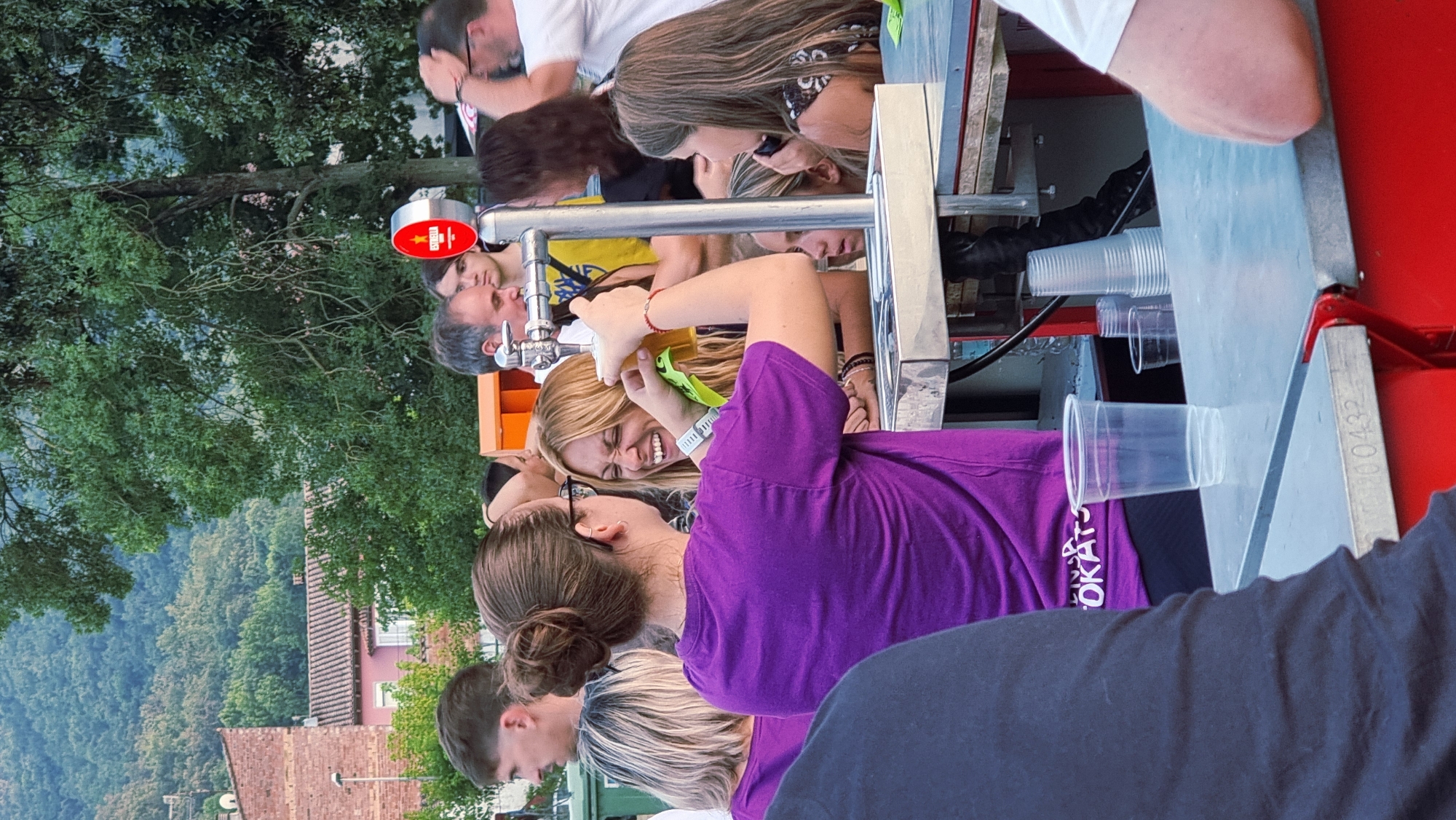 The image depicts an outdoor gathering in Sant Joan Les Fonts, Spain, with a focus on a beer tap station. In the foreground, a woman with her hair in a bun, wearing a purple t-shirt, is interacting with a laughing woman with long blonde hair who is holding a glass of beer. The blonde woman is wearing a white wristband. Behind them, a beer tap with a red "Estrella" logo is visible. Several other people are present in the background, including men and women of various ages, some of whom are holding drinks or engaged in conversation. The environment suggests a casual event, possibly a festival or outdoor party, with trees and buildings visible in the distance. The beer tap is mounted on a metallic counter, with plastic cups stacked nearby. A red structure is visible at the bottom of the frame, possibly part of the bar or stall.