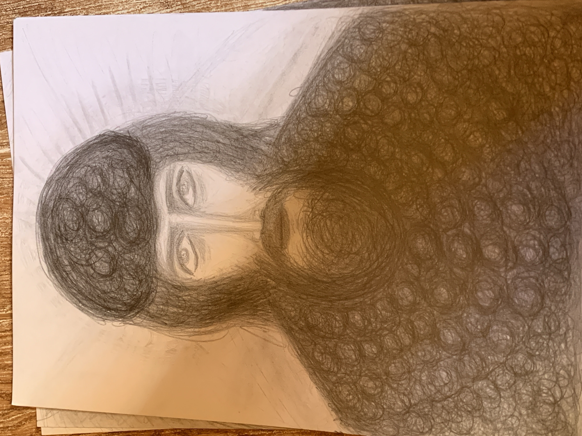 A pencil drawing on paper depicts a portrait of a person with a beard and dark, curly hair. The person is wearing a dark, textured garment. The background is rendered with faint lines radiating outwards from the subject's head, suggesting a halo or light source. The drawing is placed on a surface that appears to be a wooden tabletop.
