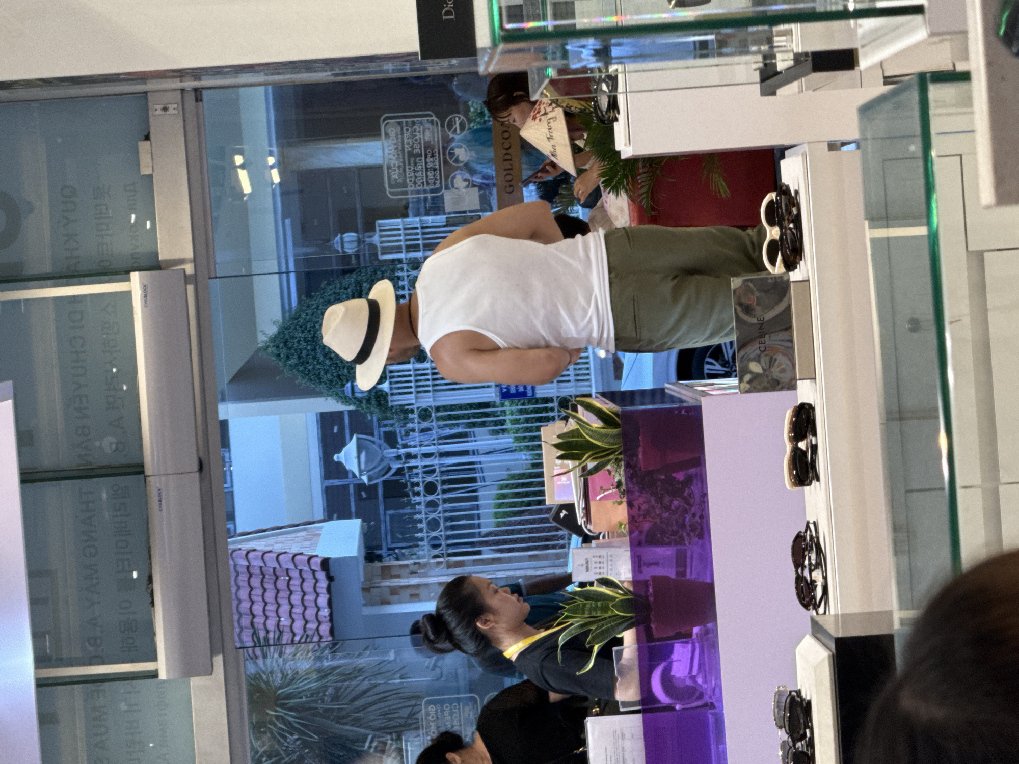 In Nha Trang Ward, Vietnam, a retail store window displays sunglasses on shelves. A man wearing a white tank top and a straw hat stands facing away from the camera, looking into the store. A woman with her hair in a bun, wearing a black t-shirt and a yellow lanyard, is seen behind a counter, looking down. Two potted snake plants are placed on the counter. Reflections in the glass show buildings and a palm tree.
